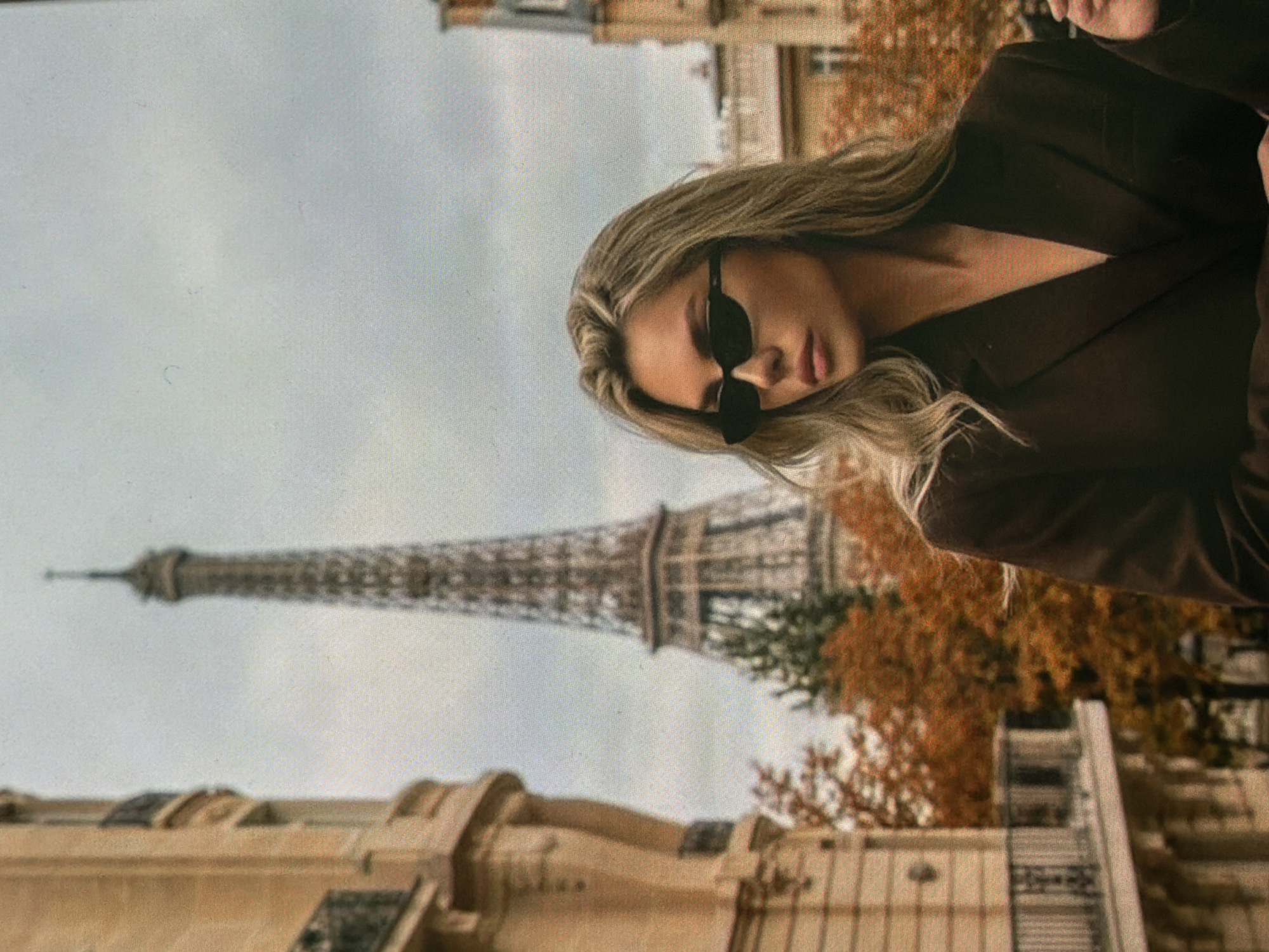 The image is an outdoor, medium shot of a blonde woman wearing sunglasses and a dark jacket, standing in front of the Eiffel Tower. The Eiffel Tower is visible in the background, along with autumnal trees and Parisian architecture. The woman is looking to her left, with her hair blowing in the wind. The sky is overcast. The location is Paris, France, not Bobrovo, Russia.