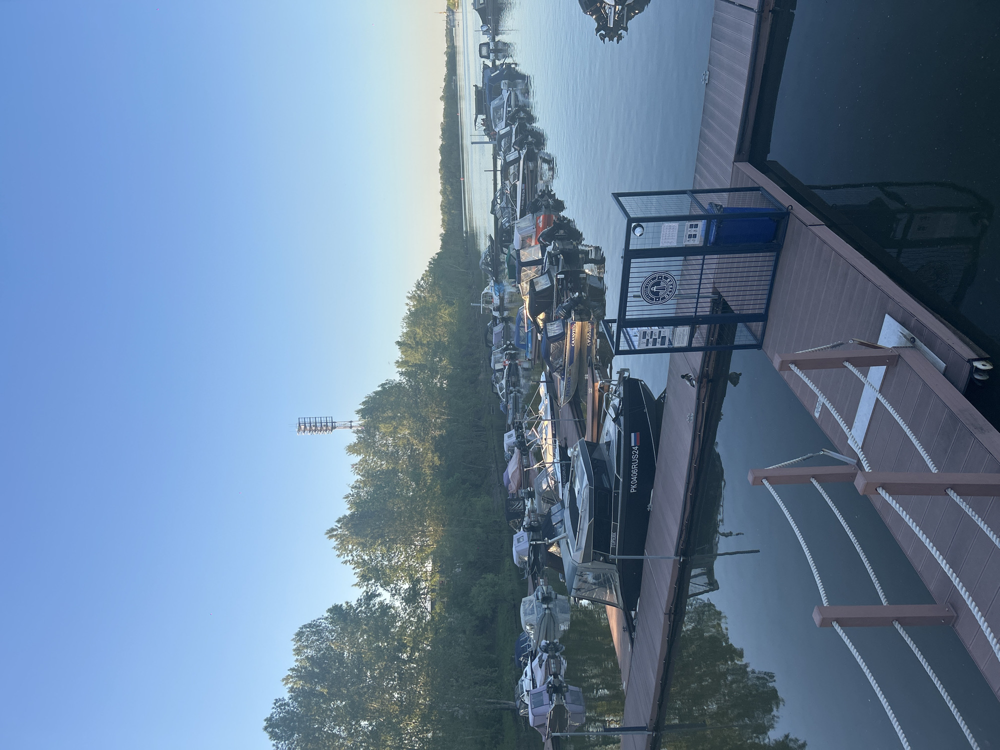 The image depicts a marina or boat dock located in Krasnoyarsk, Russia. Several boats are moored alongside a wooden dock, reflected in the calm water. The dock features a metal fence and a blue trash receptacle. Trees line the shore adjacent to the marina, and a communications tower is visible in the background. The sky is clear blue with some early morning or late afternoon light. A boat labeled "PK0406RUS24" is visible on the near side of the dock.