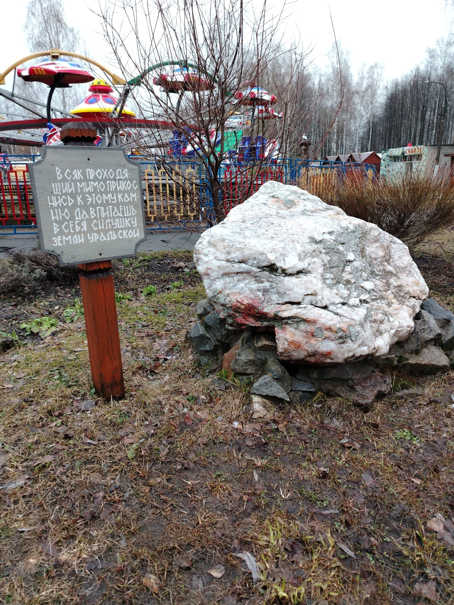 The image shows a large white and grey rock on a bed of smaller rocks, located next to a wooden sign with Russian text. The sign is positioned to the left of the rock and is supported by a wooden post. The background features a children's ride with red, yellow, and blue elements, along with bare trees and buildings. The ground is covered in dead grass and leaves, suggesting an outdoor setting in the city of Unknown, Unknown. The text on the sign reads: "ВСЯК ПРОХОДЯ ЦИЙ МИМО ПРИКОС НИСЬ К ЭТОМУ КАМ НЮ ДАВПИТАЕШЬ ОН В СЕБЯ СИЛУШКУ ЗЕМЯН УРАЛЬ СКОЙ". The presence of the children's ride indicates a park or playground environment.