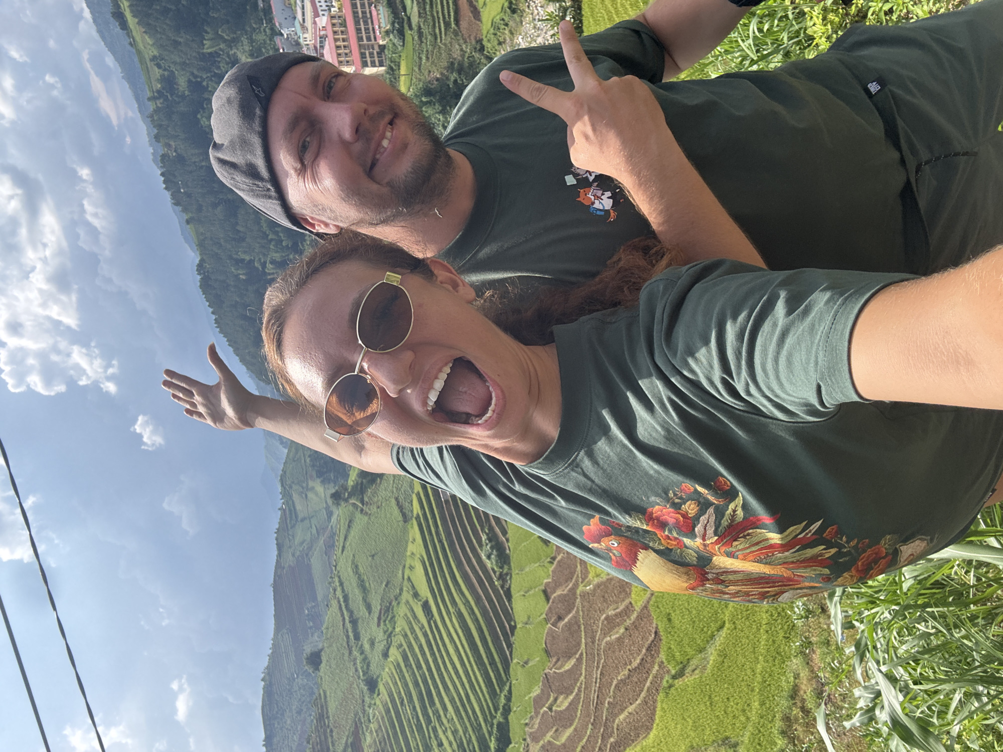 This is a vibrant, outdoor portrait featuring two smiling individuals, a man and a woman, set against a stunning backdrop of terraced rice paddies and lush mountains in Khao Mang, Vietnam. The image appears to be a selfie or a close-up photo taken from a low angle, showing them from the chest up.
The man, positioned slightly behind and to the right of the woman, has a beard and is wearing a dark grey cap and a dark green t-shirt with a small graphic on the left chest, which appears to be a stylized animal, possibly a fox. He is smiling broadly and making a peace sign with his right hand.
The woman, in the foreground, is wearing round, gold-rimmed sunglasses and a dark green t-shirt featuring a large, colorful graphic of a rooster surrounded by floral elements. Her mouth is open in an expression of excitement or laughter, and her left arm is raised as if in a gesture of joy or surprise.
The background showcases the distinctive, verdant contours of rice terraces, characteristic of mountainous regions in Vietnam. Beyond the paddies, forested mountains rise, and in the distance, parts of what appears to be a village or town with buildings are visible. The sky is bright with some scattered white clouds, indicating a clear, sunny daytime. Two dark electrical wires are visible in the upper left portion of the image. Both individuals convey strong emotions of happiness and enjoyment. There is a small, unreadable text or logo on the man's t-shirt.