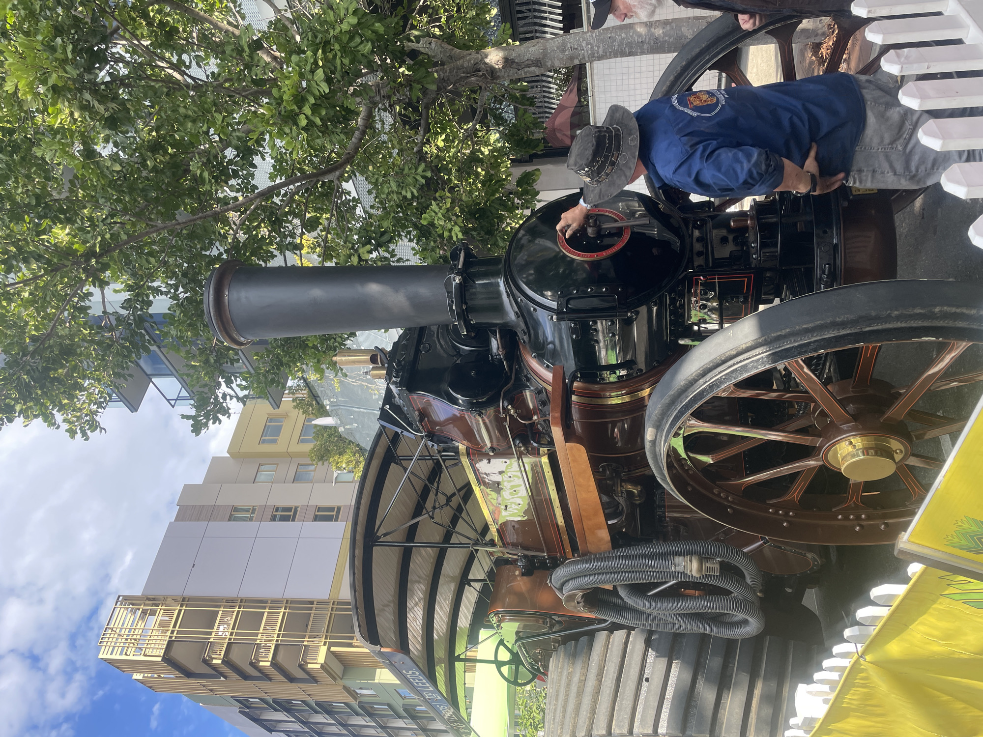 An aged, steam-powered traction engine, painted in shades of brown, black, and gold, is displayed outdoors, positioned behind a white picket fence and a yellow banner. A man, wearing a wide-brimmed hat, blue shirt with a circular emblem, and grey trousers, stands facing the engine. He is operating a circular mechanism on the front of the engine. The engine has large spoked wheels, a prominent smokestack, and a covered canopy. In the background, modern apartment buildings rise above lush green trees, suggesting an urban setting. The blue sky is visible with some scattered clouds. The overall scene captures a juxtaposition of historical machinery and contemporary city architecture.