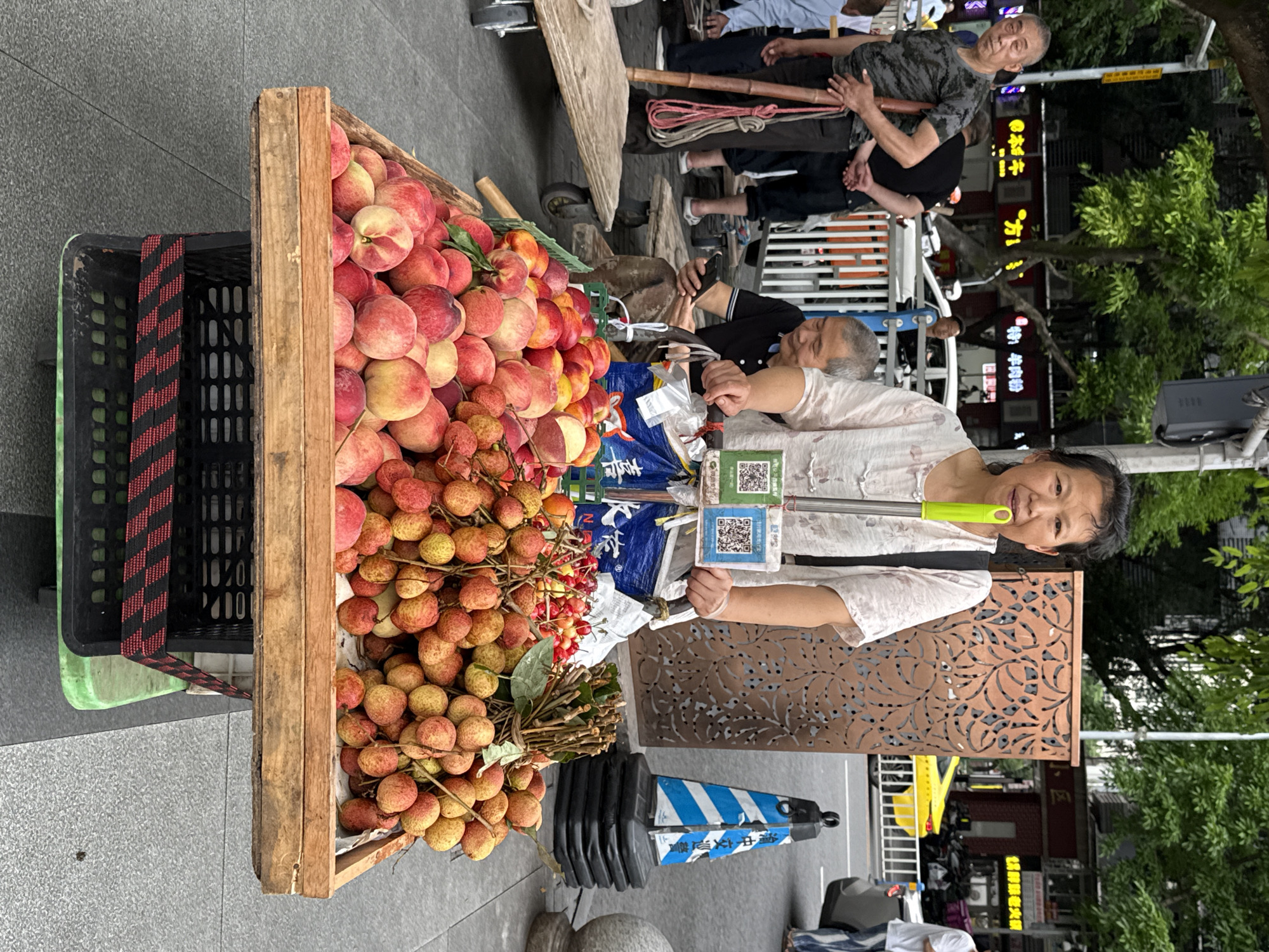 A woman stands next to a wooden cart filled with peaches and lychees on a sidewalk in Yuzhong District, China. The cart is laden with a large quantity of fresh produce. The woman, dressed in a white blouse, is smiling and appears to be selling the fruit. Other people are in the background, some seated. Additional elements include a black plastic crate, a decorative screen, a traffic marker, and the urban environment with buildings and greenery.