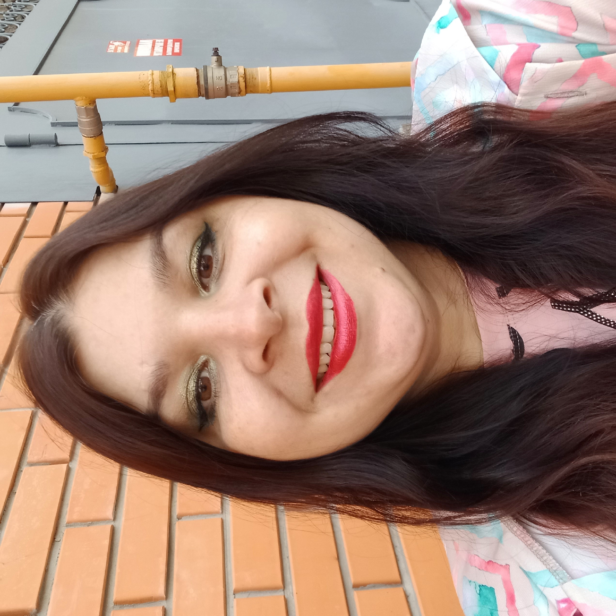 The image is a medium shot, taken outdoors, of a woman with dark brown hair and fair skin. She is smiling and looking at the camera. Her makeup includes green eyeshadow and red lipstick. She is wearing a pink shirt with a patterned jacket over it.
Behind the woman, there is a brick wall on the left and a grey metal gate on the right. A yellow pipe, possibly part of a gas line, runs vertically on the right side of the image, attached to the grey gate. The background suggests an urban environment, consistent with a location in Kropyvnytskyi, Ukraine, where gas lines are common utilities installed on the exterior of buildings. The overall lighting and setting indicate it is daytime.