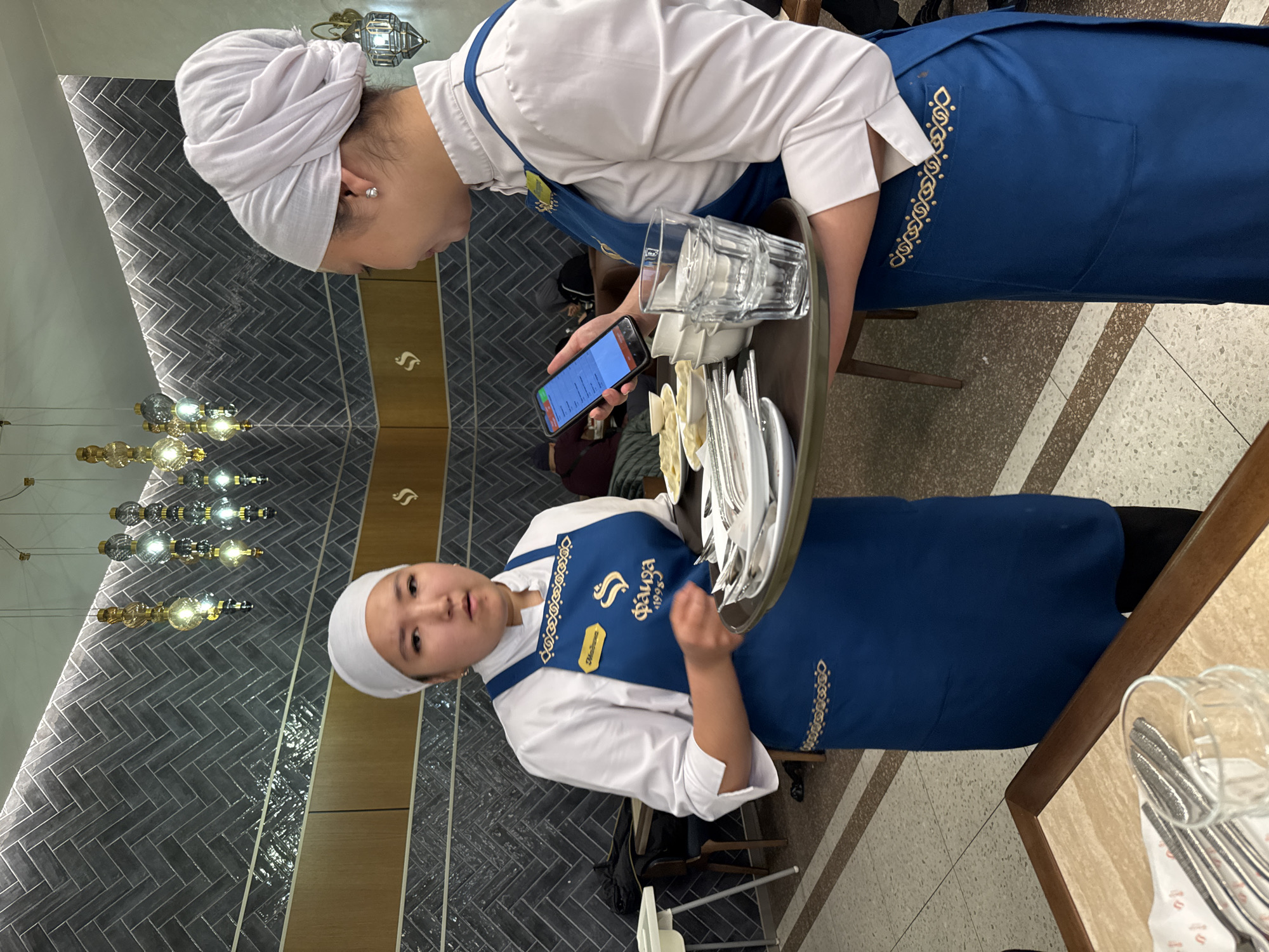 Two young women, likely waitstaff, are depicted in an indoor restaurant or cafe setting in Bishkek City, Kyrgyzstan. Both are wearing white long-sleeved shirts, blue aprons with intricate gold embroidery, and white head coverings resembling turbans or wraps.
The woman on the right side of the image (from the viewer's perspective) is holding a dark serving tray laden with white plates, silverware (forks, knives, spoons), small white bowls, and clear drinking glasses. She is looking down towards the tray. The woman on the left is holding a smartphone in her hand, which is positioned above the tray, and she appears to be reviewing content on its screen. Her gaze is directed towards the phone. The activity suggests they are either preparing an order for service or checking details related to the items on the tray using the phone.
Notable details include the uniform design, specifically the gold embroidery on the blue aprons, which features traditional-looking patterns. On the apron of the woman on the right, the large text "Файза" (Fayza) and "1995" are visible, along with a yellow nametag that is partially obscured but seems to read "Мадина" or similar. The smartphone screen displays what appears to be an application, possibly an order system or menu, with some red accents at the top.
The background features a dark grey tiled wall with a herringbone pattern, accented by horizontal strips of a light brown or wooden material and reflective vertical panels. Modern hanging light fixtures with multiple exposed bulbs are visible. The flooring is a light-colored speckled material, possibly terrazzo. The overall ambiance is that of a contemporary dining establishment. No specific time of day or outdoor weather is discernible.