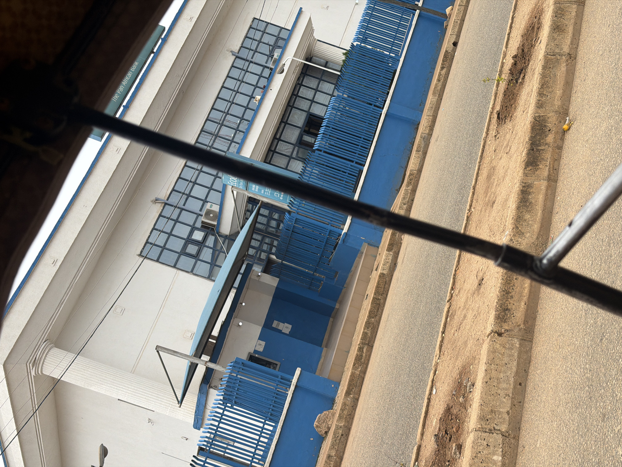 The image shows a street scene in Bukuru, Nigeria, featuring a building with "The Pan African Bank" signage. The bank is a two-story structure with a white facade, large blue-tinted windows, and prominent white columns. A blue metal fence runs along the front of the property, separating it from the road. The road is paved and shows signs of wear, with a concrete curb and a sandy median where a small sapling is planted. A dead yellow bird lies on the side of the road near the curb. The perspective suggests the photo was taken from inside a vehicle or through a window, with dark framing elements visible in the foreground.