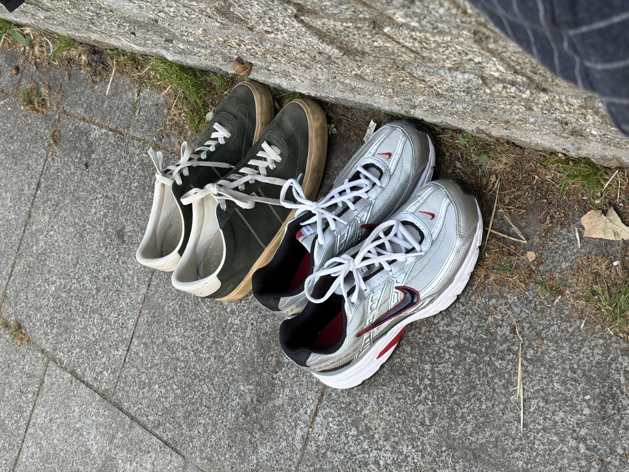 The image shows two pairs of shoes placed near a stone ledge on a gray concrete surface. The first pair of shoes are green suede sneakers with white laces and a tan sole. The second pair of shoes are silver athletic sneakers with red and black accents and white soles. The laces of both pairs are tied together. The location appears to be outdoors, possibly in Belgrade, Serbia. The background includes grass, some dry leaves, and a section of the stone ledge.