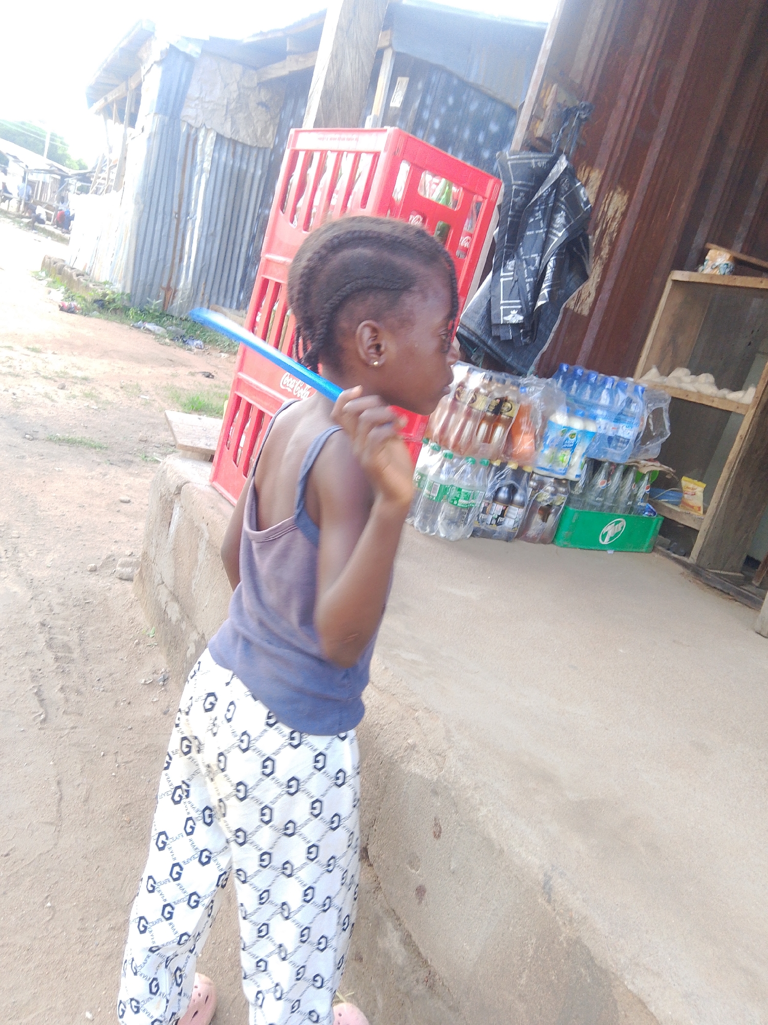 In Jalingo, Nigeria, a young Black girl stands in profile, looking to her right. She has braided hair and wears a grey tank top and white pants patterned with black "G" logos and other symbols. A blue plastic object is held near her face, its purpose unclear. Behind her, a red Coca-Cola crate is visible, along with various bottles of drinks in plastic packaging, some of which are branded. A dark, possibly black plastic bag hangs from a structure behind the drinks. The background shows a building with corrugated metal siding, suggesting an outdoor market or roadside stall. The ground is dusty earth. The lighting indicates it is daytime, with the sun casting a bright light, possibly obscuring some details. The scene appears to be a casual, everyday moment in a bustling environment.