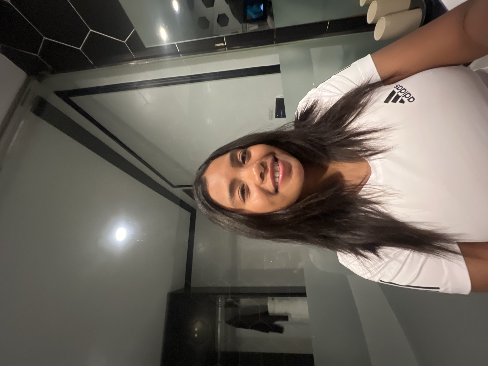 In a dimly lit room, a young woman with long dark hair and brown skin smiles at the camera. She is wearing a white Adidas t-shirt with black stripes on the sleeves and the Adidas logo on the chest. The background appears to be a modern bathroom with glass partitions, black and white hexagonal tiles, and a ceiling light fixture. There are reflections in the glass, showing what might be a closet and a television screen. The scene is likely in Hà Nội, Vietnam, given the context.
