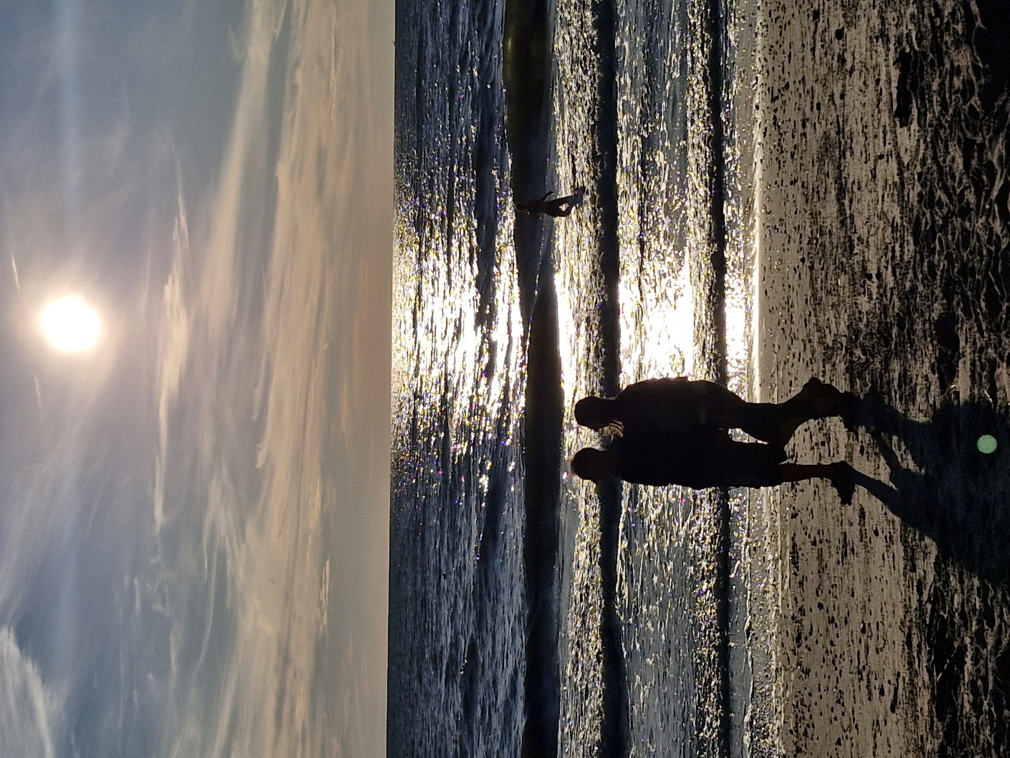 A vertical, full shot captures a serene sunset over the Baltic Sea in Karklė, Lithuania. The sun, a bright white orb, hangs high in the hazy sky, casting a shimmering path of light across the choppy water. Wispy clouds streak across the sky, painted in soft hues of peach and blue. On the sandy shore in the foreground, two figures walk away from the camera, their silhouettes dark against the illuminated sea. They appear to be a couple, walking side-by-side. Further out in the water, a lone surfer rides a wave, a small, dark figure against the vast expanse of sea and sky. The beach is sandy and slightly textured, with subtle footprints visible. The overall impression is one of tranquility and natural beauty.