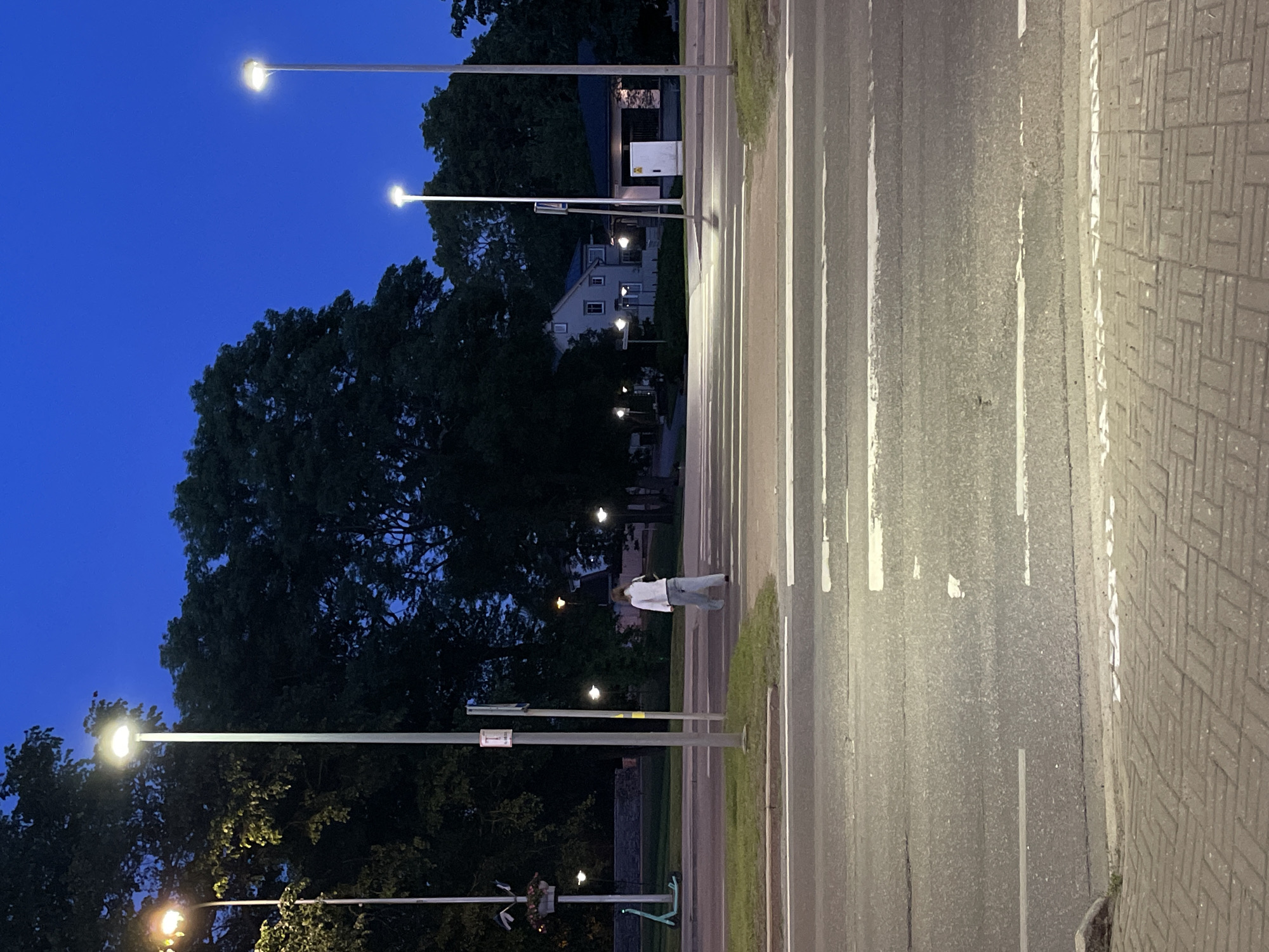 The image captures a street scene in Tallinn, Estonia, at dusk. The sky is a deep twilight blue. A person, wearing a light-colored jacket and loose-fitting pants, is walking across a pedestrian crossing on a road. The crossing is marked with white stripes. Streetlights illuminate the area, casting light on the road and the surrounding trees. In the background, houses and more streetlights are visible. The foreground shows a brick-patterned sidewalk. A scooter is parked on the grass to the left of the road. The overall impression is one of a quiet evening in a residential or mixed-use area of the city.