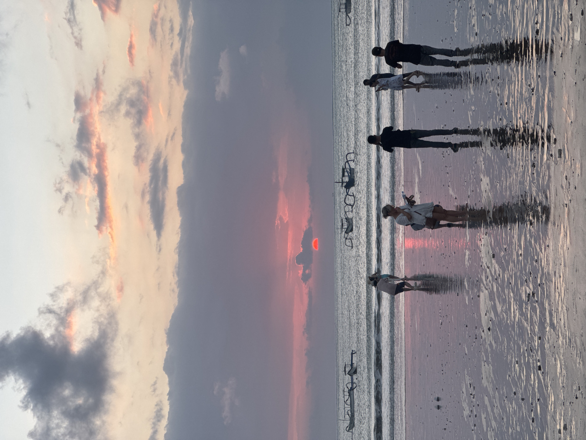 The image presents a picturesque sunset scene on a beach, likely in Kuta Selatan, Indonesia. The sky dominates the upper and middle sections, filled with a dramatic interplay of dark grey clouds and clouds brilliantly lit by the setting sun in shades of orange, pink, and soft red. The sun itself is visible as a distinct, vibrant red-orange disc, partially obscured by clouds, positioned low on the horizon.
Below the colorful sky, the ocean stretches to the horizon with gentle, reflective waves. The foreground features an expansive stretch of wet sand, which beautifully mirrors the sky's colors and the figures standing upon it, creating long, dark reflections.
Several individuals are present on the beach, enjoying the evening. On the left side, a person in a light-colored top and shorts, wearing a hat, stands near the water's edge. Another individual in light attire is positioned further to the right. Towards the right side of the frame, two figures are visible as distinct dark silhouettes against the reflective wet sand, possibly walking or standing. A fifth person is partially visible behind these two.
Scattered across the water, several dark, elongated boat-like structures, reminiscent of traditional Indonesian fishing boats (jukung), appear as silhouettes, enhancing the local character of the scene. The overall activity involves people leisurely strolling or standing on the beach during this beautiful sunset. The scene conveys a sense of calm and natural beauty, characteristic of a tropical beach at dusk. There is no visible text in the image.