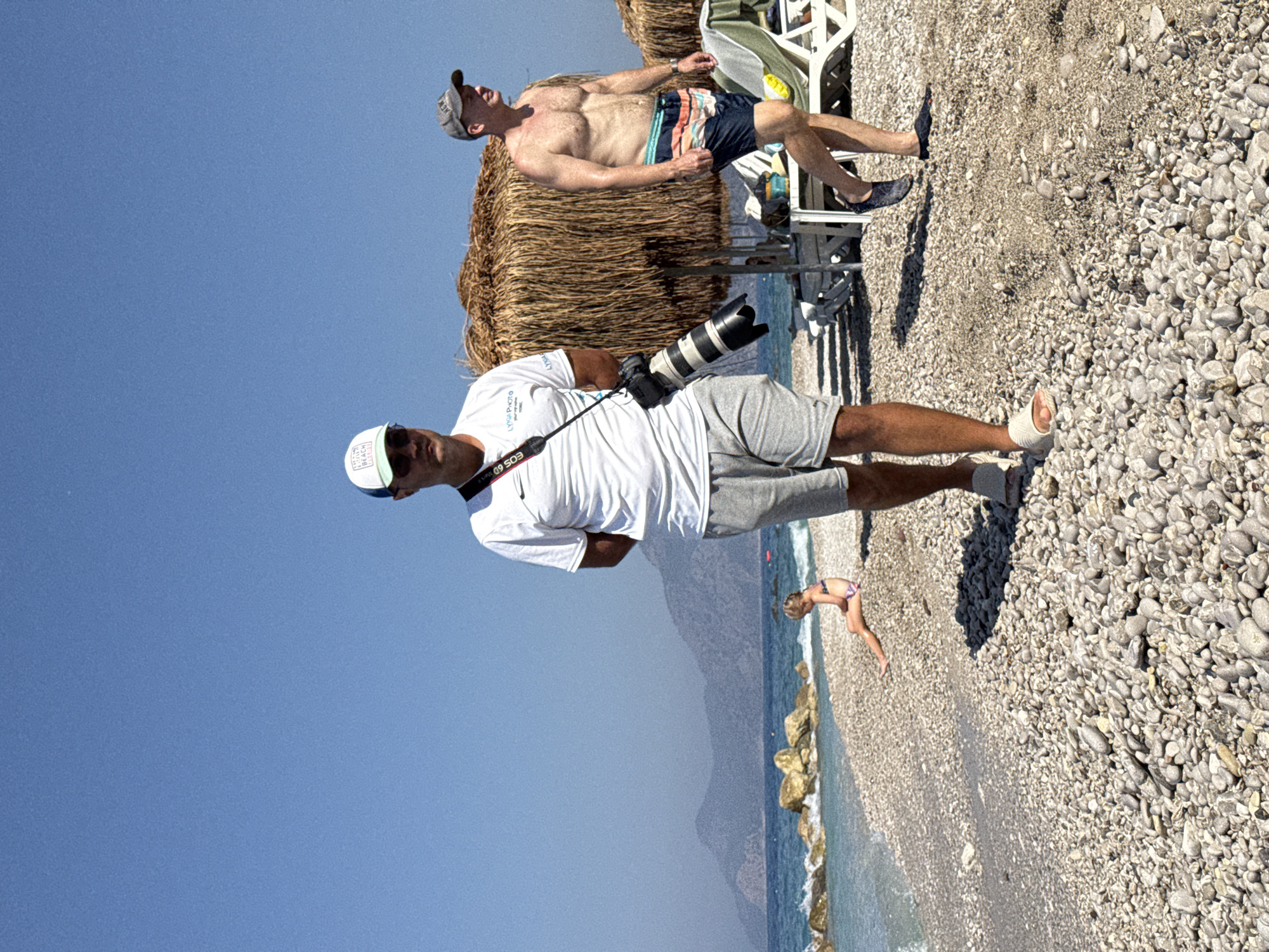 In this outdoor, eye-level shot, two men are the primary subjects, with a child visible in the background near the water. The scene is a sun-drenched beach, characterized by its extensive pebble shore and the clear blue sky overhead. The time of day appears to be midday, given the bright sunlight and the absence of long shadows.
The man in the foreground is wearing a white t-shirt and grey shorts, and is holding a professional camera with a large lens. He is wearing a white baseball cap and sunglasses. The second man, positioned behind and to the left of the foreground subject, is shirtless and wearing striped swim trunks. He is wearing a baseball cap and is reclining on a straw-thatched structure, possibly a beach umbrella or shelter. His gaze is directed upwards.
In the background, a small child in a bathing suit is playing near the turquoise water. The beach is lined with rocks and the sea stretches out towards a distant, hazy mountain range. The overall atmosphere suggests a relaxed, vacation setting.
The text "EOS 6D" is visible on the camera strap in the foreground, indicating the camera model. The cap worn by the foreground subject has text, but it is partially obscured and difficult to read.
The location is Fethiye, Turkey, which is consistent with the coastal scenery and the Mediterranean climate implied by the bright sun and clear water. The activity is people enjoying a day at the beach, with one person photographing and others relaxing or playing.
