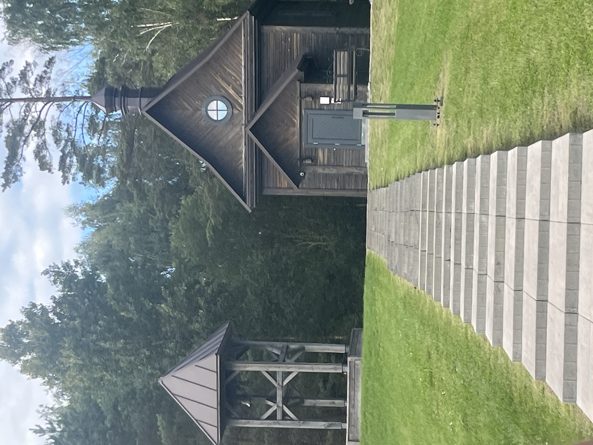 The image shows a path of light grey stone steps leading up a grassy incline towards a rustic wooden building, possibly a chapel or a small church, with a dark metal roof and a small circular window. To the left of the building and slightly further back, there is a wooden bell tower with a dark metal roof. The structures are set against a backdrop of dense green trees under a cloudy sky. A dark grey modern lamp post is visible near the steps. There are no people visible in the image. The scene appears to be located in a rural or natural setting, consistent with the Каменскі сельскі Савет area of Belarus, which is known for its natural landscapes and historical sites.