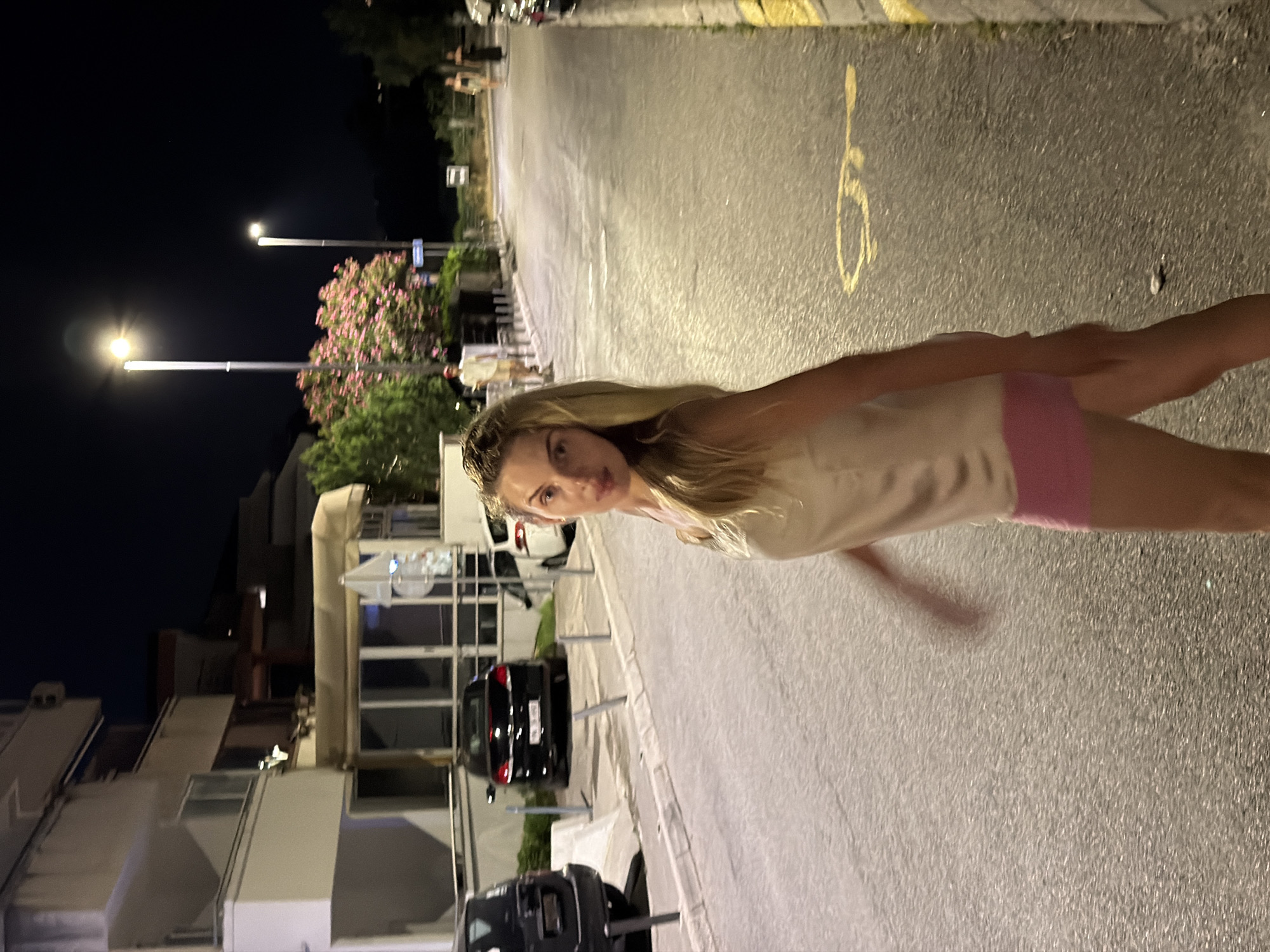 In Budva, Montenegro, a young woman with blonde hair is captured at night, walking across a street. She is wearing a light-colored camisole and pink shorts. The street is paved and appears to be a public road with parked cars and buildings lining one side. Streetlights illuminate the scene, casting a warm glow on the asphalt and pavement. In the background, several other individuals are visible, also walking along the street. The overall atmosphere is that of a quiet, urban evening in a coastal town.