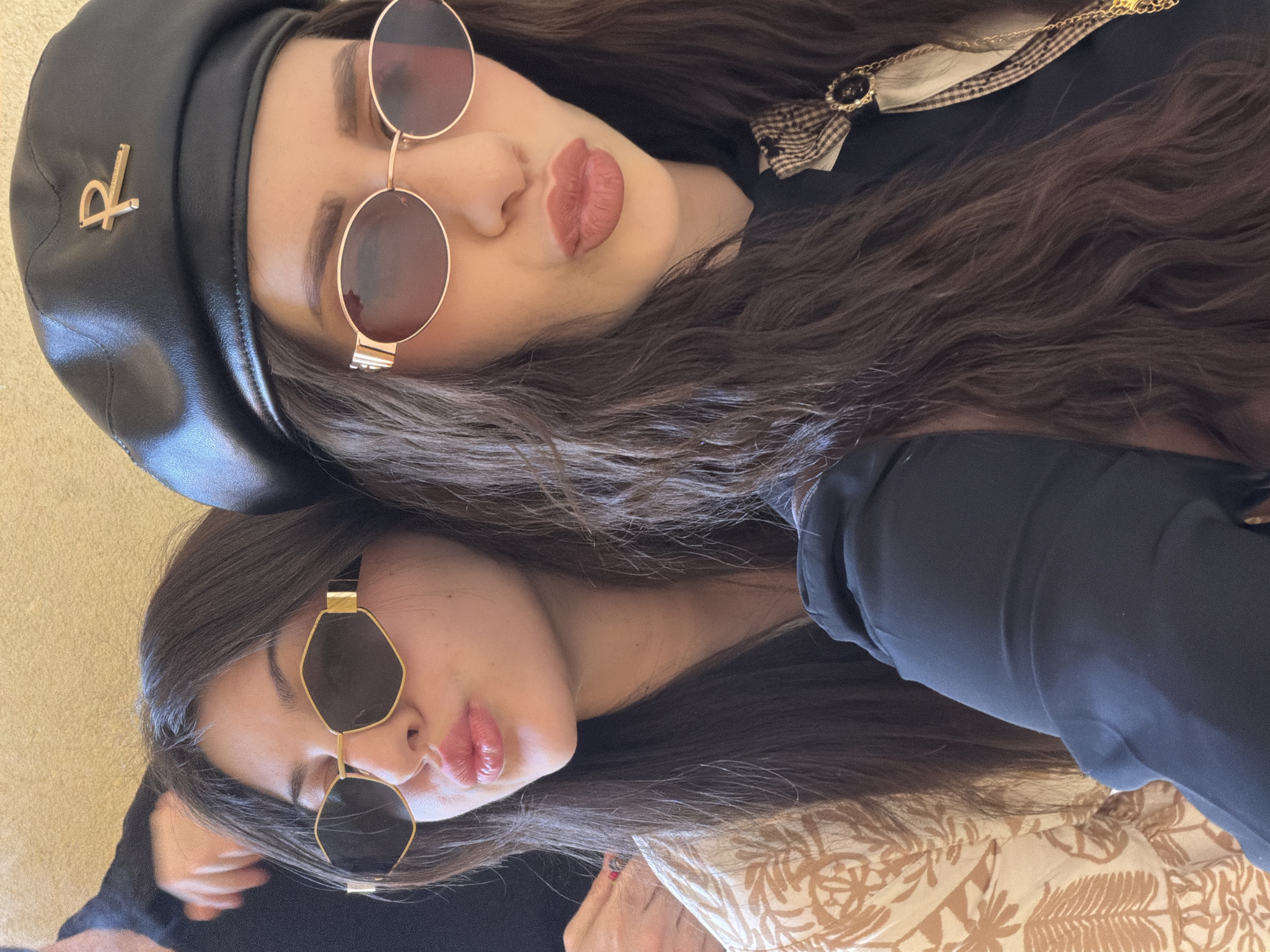 Two women are lying down next to each other. The woman at the top of the image wears a black leather beret with a gold "R" logo and round-framed sunglasses with reddish lenses. She is pouting her lips. The woman beneath her wears geometric, dark sunglasses and is also pouting. Both women have dark hair. They are wearing black clothing. A patterned beige and brown blanket or sheet is visible in the lower part of the image. The location is not identifiable as Milan, Italy, based on the visual information provided.