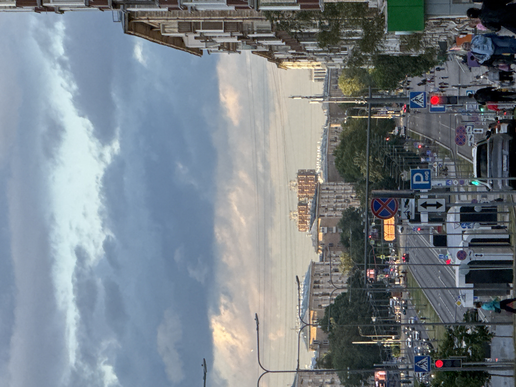 The image depicts an urban street scene in an unknown city. The perspective is looking down a wide road lined with multi-story buildings on the right and trees and lower buildings on the left. The sky is partly cloudy with a mix of dark gray and lighter, possibly sunlit, clouds.
Vehicles are visible on the road, suggesting ongoing traffic. Traffic lights and various street signs, including parking signs, pedestrian crossing signs, and what appears to be a "no stopping" sign, are present along the street. Several street lamps are also visible.
People are walking on the sidewalks, and some are near crosswalks. The buildings on the right are tall and have many windows and balconies. The buildings on the left appear to be of a similar architectural style but are partially obscured by trees. In the distance, more buildings and structures are visible, including what looks like a construction site or a building with a distinct modular design. The overall impression is that of a bustling urban environment during daylight, possibly late afternoon or early evening, given the lighting conditions.