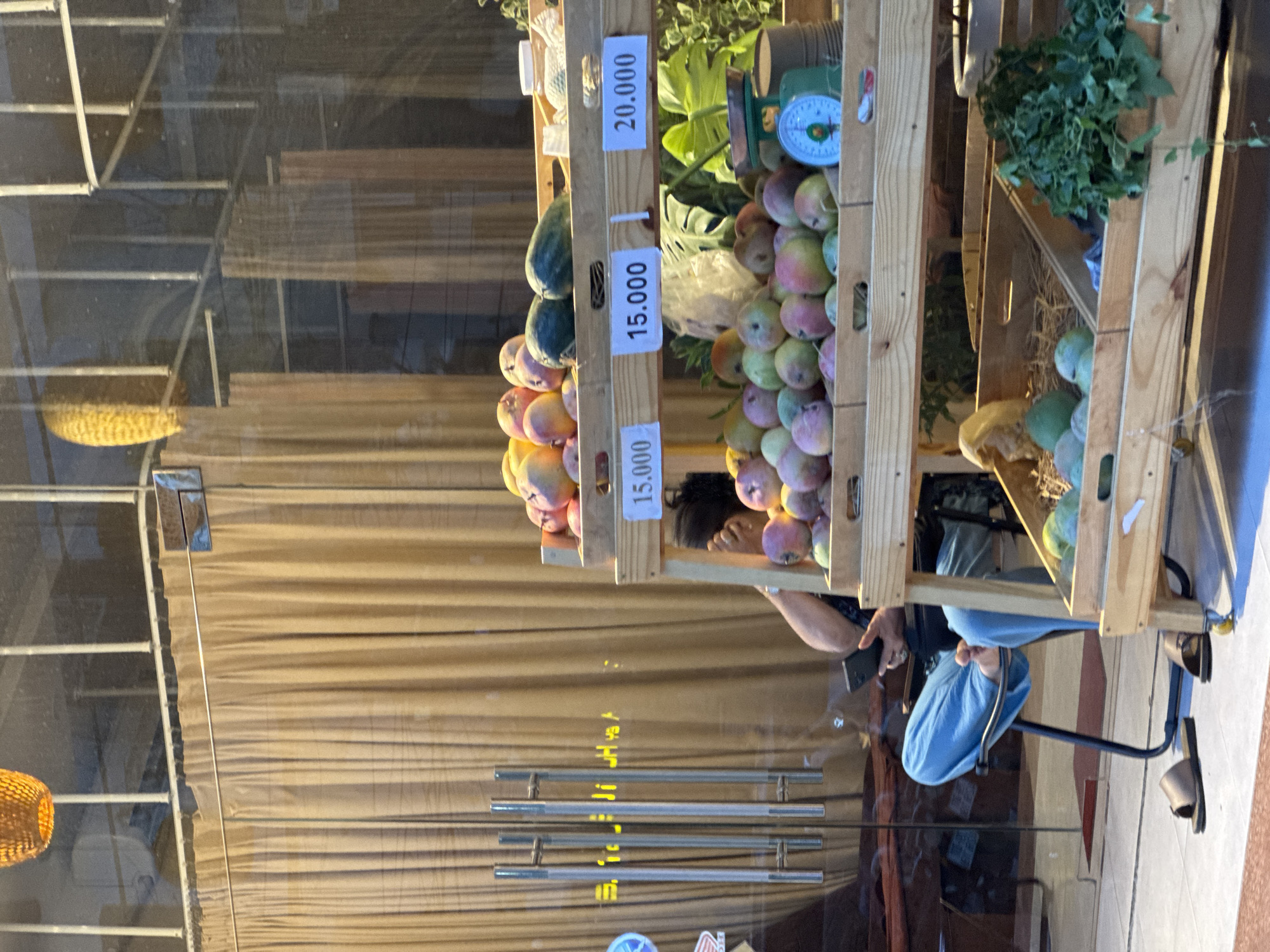 A street view shows a fruit stand with several wooden shelves displaying fruits. The first shelf contains mangoes and two green fruits, possibly papayas or watermelons, along with a jar. Price tags are visible: "15.000" and "20.000". The second shelf also displays mangoes, some of which appear to be smaller and more varied in color. A vintage green scale with a blue dial and a metal tin are present on this shelf. The bottom shelf has a wooden crate containing more fruits and some green leafy vegetables. A person is seated on a chair to the left, wearing blue pants and a dark shirt, holding a black smartphone. There are two pairs of sandals on the ground near the chair. The background features a beaded curtain and glass doors with reflective surfaces, showing the interior of a building and some reflections of the street. A woven orange light fixture hangs from the ceiling. The setting is likely a market or shop in Bac Nha Trang Ward, Vietnam.
