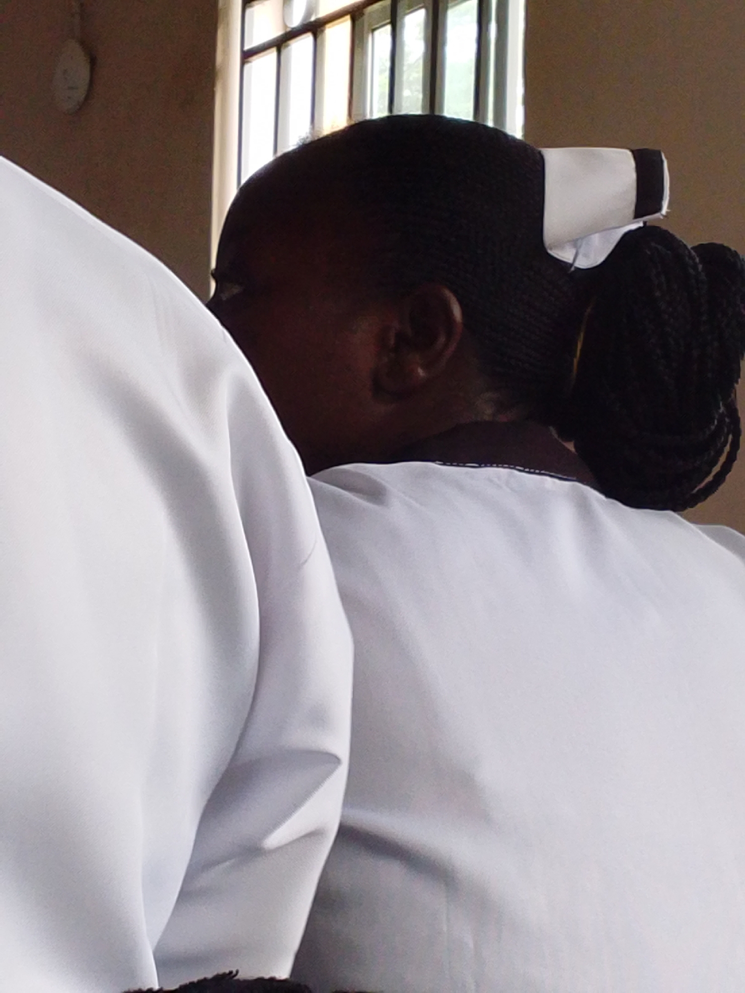 The image shows the back of two people wearing white garments, likely uniforms. The person in the foreground is the focus, their head and shoulders visible. They have dark, braided hair styled in a bun and are wearing a white cap with a black band. Their skin tone is dark. The person next to them, to the left, is also wearing a white garment and their shoulder is visible. The scene appears to be indoors, with a wall and a window with several panes in the background. The light coming from the window suggests it could be daytime. There is no text visible in the image.