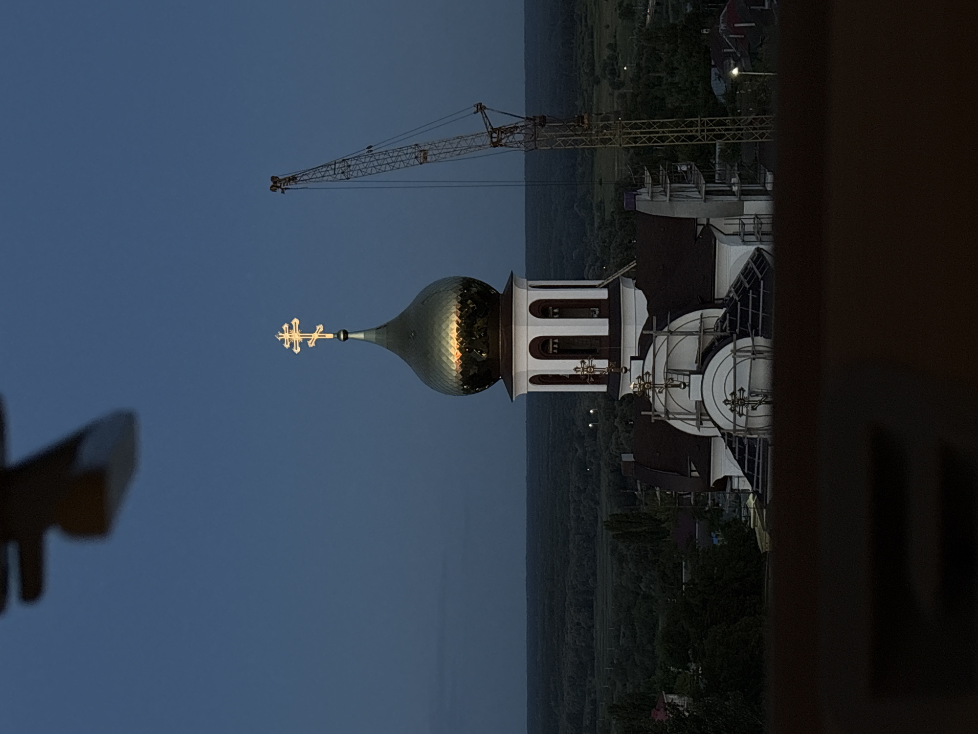 The image shows a golden dome of a church with a cross on top, illuminated by the last rays of sunlight. A construction crane is visible to the right of the church, suggesting ongoing construction or renovation. The church is situated in Bobrov, Russia, as indicated by the landscape and architectural style, which is typical of Russian Orthodox churches. The surrounding area appears to be a mix of trees and buildings, with a dark, clear sky overhead. The overall impression is one of serenity and architectural beauty, juxtaposed with the ongoing human endeavor of construction.