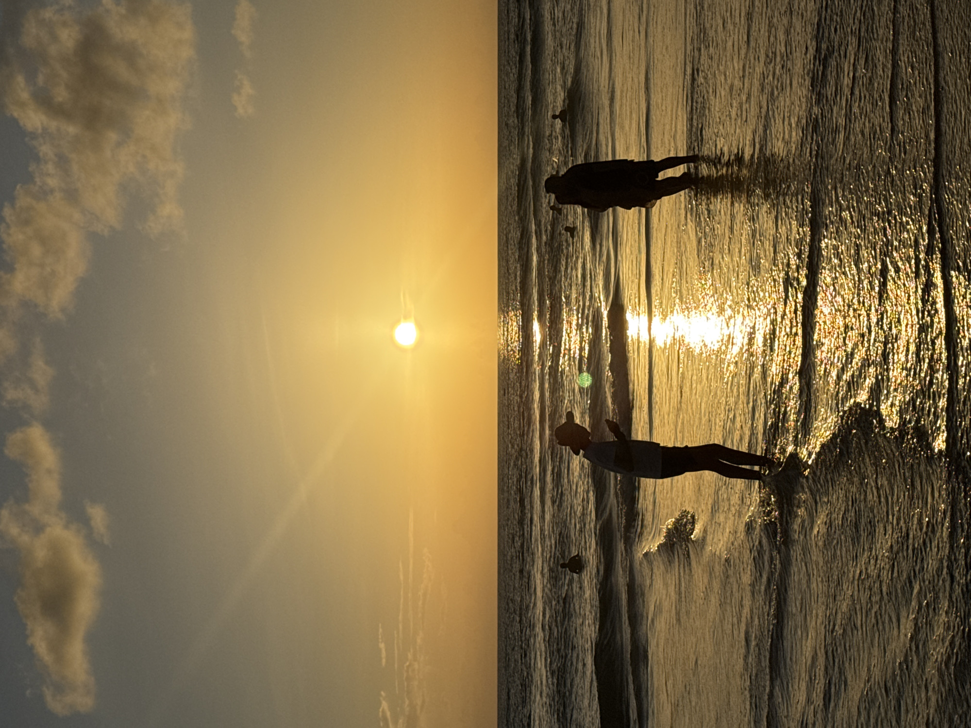 The image shows a silhouetted sunset over Karon Beach, Thailand. Two individuals stand in the shallow water, their figures dark against the bright, golden glow of the sun low on the horizon. The sun's reflection shimmers on the gentle waves that lap at their feet. In the background, the ocean stretches out to meet the sky, with a few scattered clouds tinged with the sunset's hues. Other people are visible as distant silhouettes in the water. The overall scene captures a serene and tranquil moment on the beach.