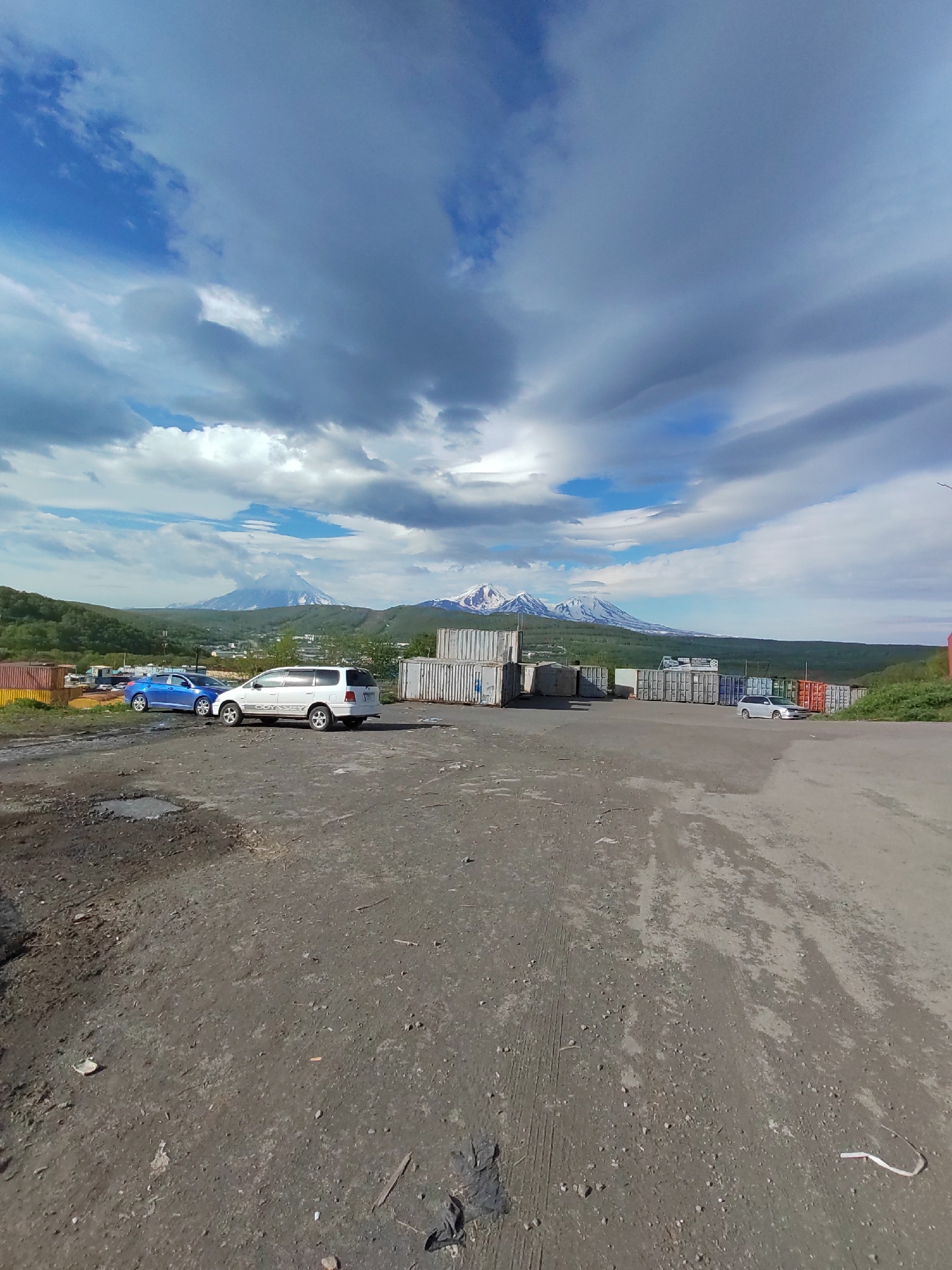 The image captures a wide-angle view of an industrial or storage area, likely on the outskirts of Petropavlovsk-Kamchatsky, Russia. In the foreground, a dirt and gravel lot is visible, with some scattered debris. Two passenger vehicles are parked on the lot: a blue sedan and a white minivan. Behind the vehicles, a line of shipping containers is arranged, indicating a storage or transport facility. In the background, a lush green hillside with scattered trees forms the middle ground, leading to a majestic mountain range with snow-capped peaks under a dramatic sky filled with clouds. The overall scene suggests a working industrial area against a backdrop of natural, mountainous beauty characteristic of the Kamchatka region.