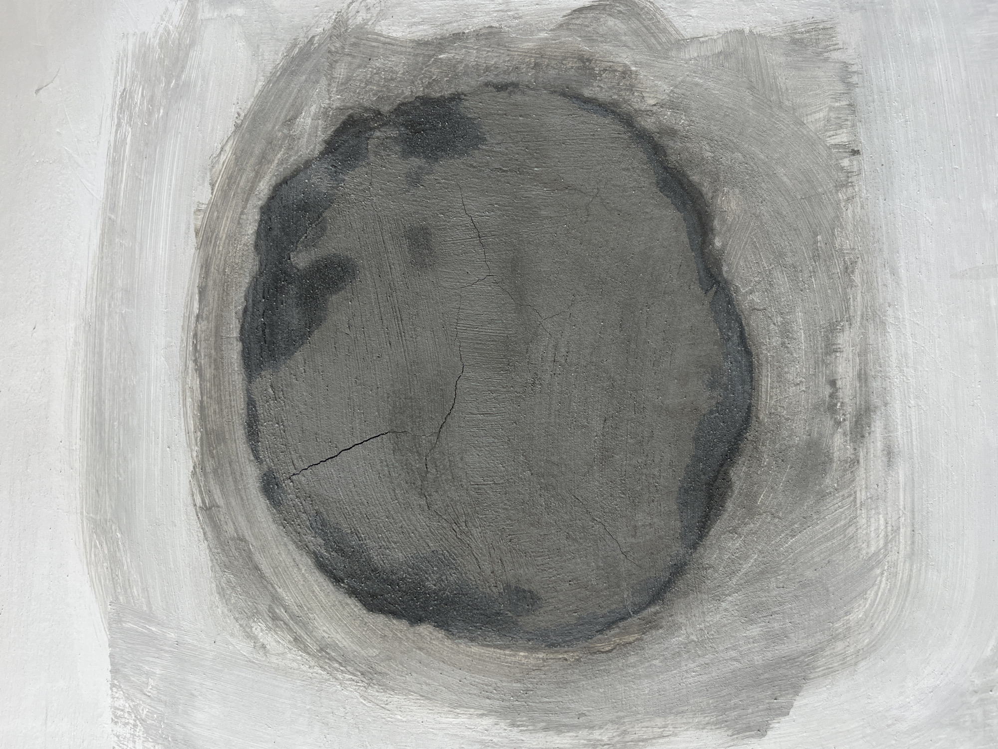 The image shows a close-up view of a weathered, oval-shaped patch on a wall, likely made of concrete or plaster. The patch is a darker shade of gray compared to the surrounding lighter gray and white painted surface. The texture of the patch is rough and shows signs of age and wear, with visible cracks and inconsistencies in the material. The outer edges of the patch are blurred and smudged, suggesting it may be a repair or a deliberate artistic application. The overall impression is one of a textured, abstract surface with a focus on the interplay of light and shadow on the rough material. There are no people, objects, or actions depicted in the image. The image does not provide any information about its location in Kyiv, Ukraine.