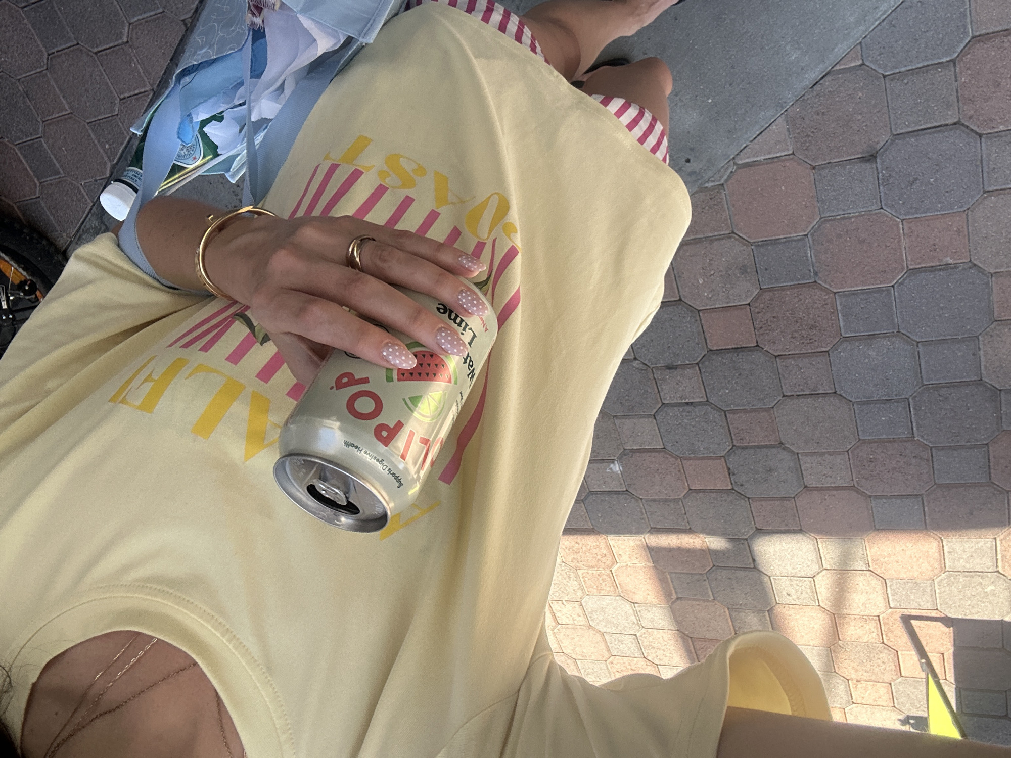 Here is a detailed description of the image:
A person wearing a yellow t-shirt is holding a can of Olipop Watermelon Lime drink. The individual is also wearing a gold bracelet and a gold ring. The person is sitting outdoors on a brick surface.