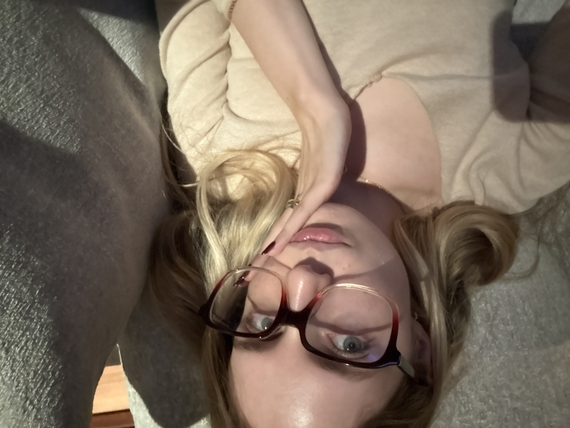 A young adult Caucasian female is lying down on a grey sofa or cushioned surface. She wears glasses with brown frames, and a light beige top. Her blonde hair is partially styled in a bun. Her right hand is positioned near her forehead. The location cannot be determined from the image.