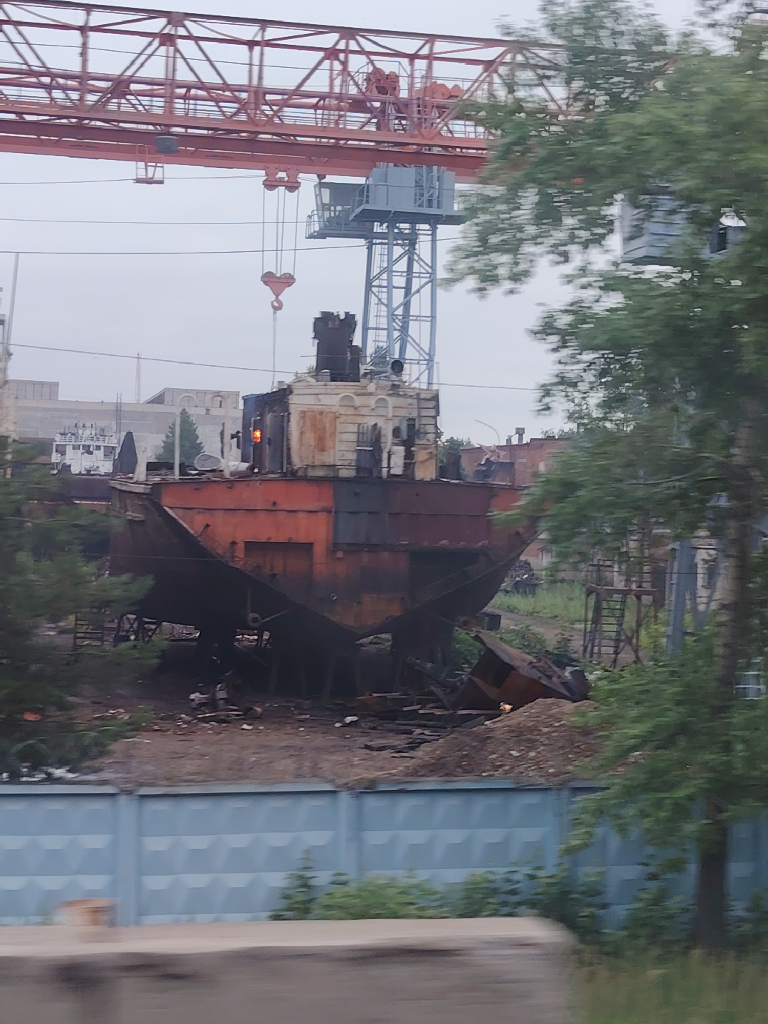 The image depicts a section of a shipyard or industrial area in Chaikovsky, Russia. In the foreground, a large, rusted ship hull is prominently displayed, sitting on land surrounded by debris and overgrown vegetation. The ship appears derelict, with sections of its structure exposed and weathered.
Above and behind the ship, a large red industrial crane dominates the upper portion of the frame, indicating ongoing or past construction or demolition activities. Further in the background, various industrial buildings and structures are visible, some appearing modernized and others in disrepair. The overall atmosphere is one of industrial activity and decay. The presence of the crane and the large ship hull strongly suggests a port, shipyard, or heavy industry zone. The faint outline of other vessels in the distance and the general industrial landscape reinforce this interpretation.