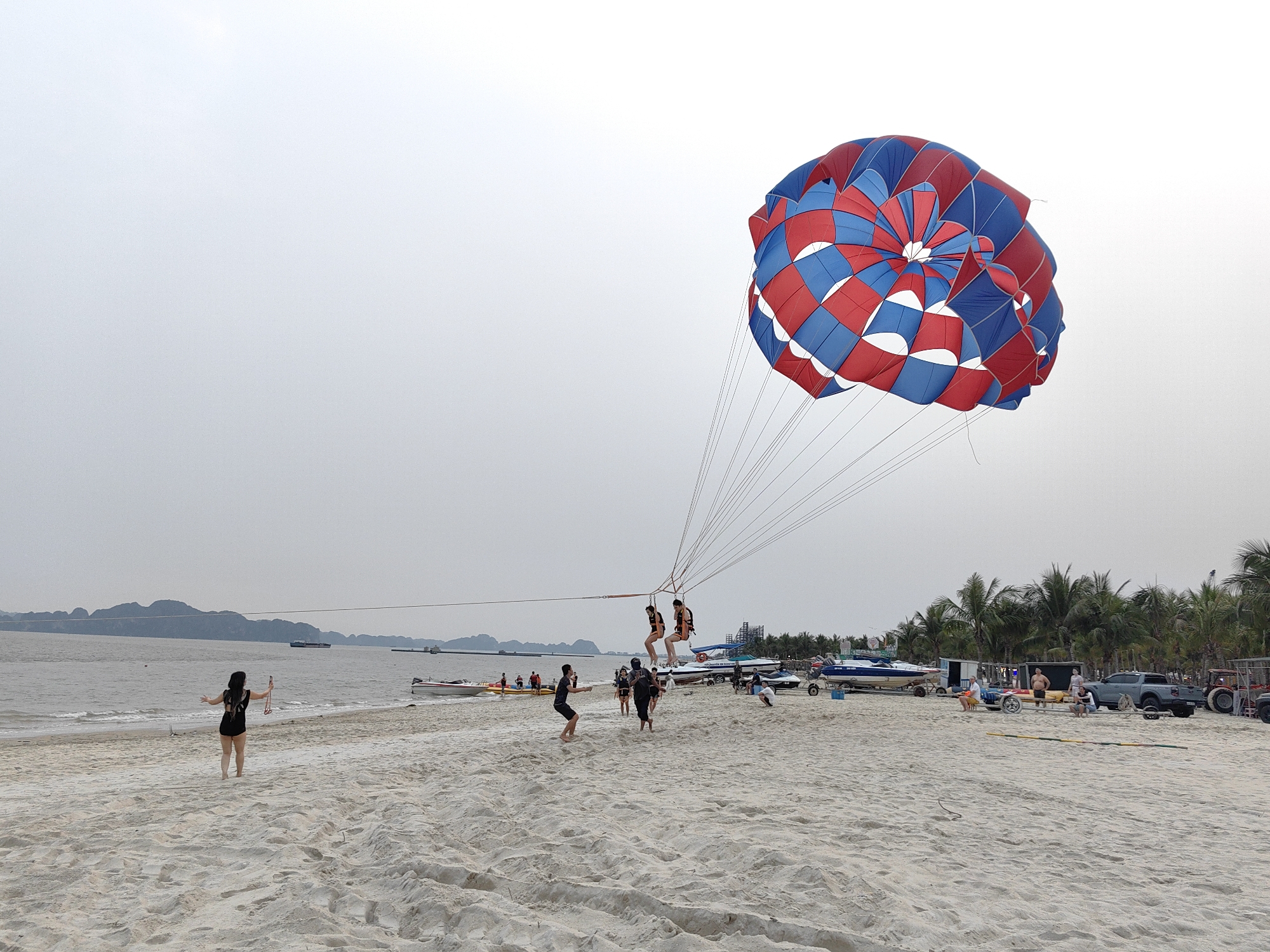 The image captures a beach scene in Bai Chay Ward, Vietnam, with a primarily overcast sky. In the center of the frame, two people are suspended in the air via a parasail that is red, white, and blue. The parasail is connected to a group of people on the ground by multiple cords. The beach itself has a light-colored sand and shows footprints. There is one woman standing alone in a bathing suit near the water. There are several boats and other watercraft positioned on the beach. The background includes palm trees, other foliage, and a mountain range in the distance. Several people can be seen standing and sitting on the beach in the area around the boats and foliage. There is also a pickup truck and a tractor on the beach to the right.