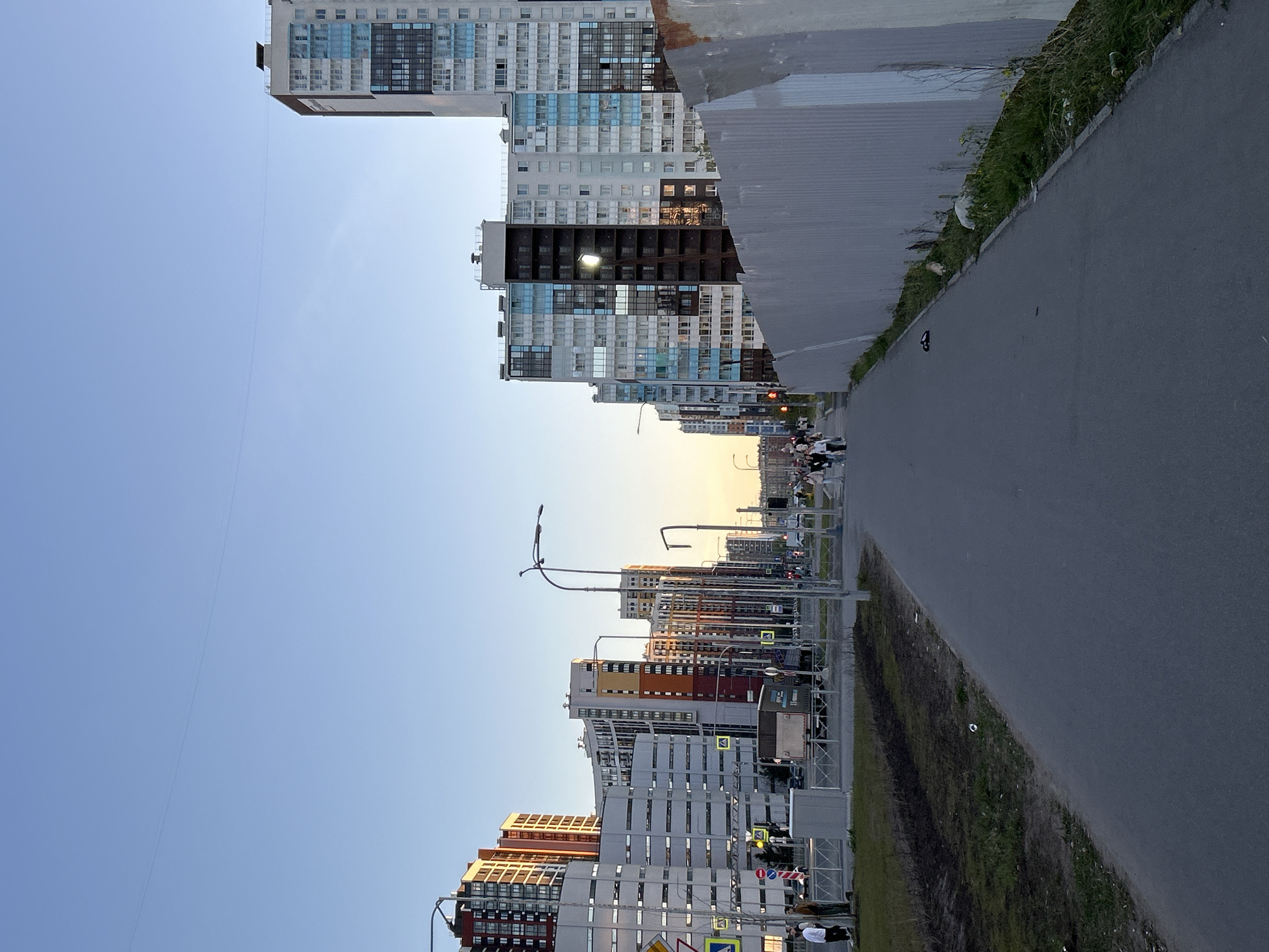 The image depicts a street scene in Saint Petersburg, Russia. High-rise apartment buildings line the left side of the street, varying in color and architectural style. A paved road runs along the right, flanked by a grassy verge. Streetlights are visible along the road, and several people are walking in the distance. The sky is clear blue, suggesting either early morning or late afternoon. The lighting indicates it is late afternoon or early morning. The scene is urban, with typical infrastructure elements.