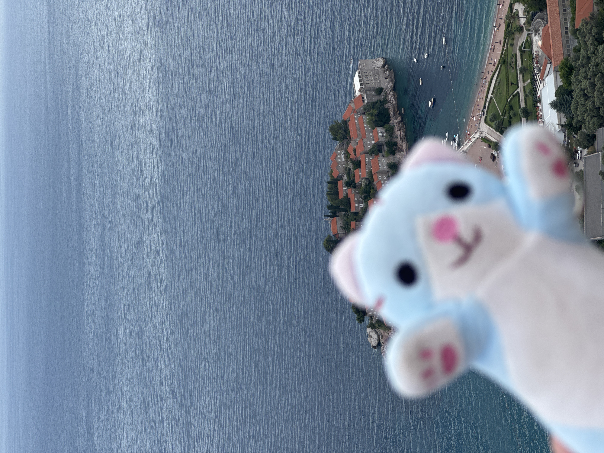 This media file shows a view from above of Sveti Stefan, Montenegro, with a blurred plush toy in the foreground. The plush toy is light blue with white paws and has pink accents on its ears and paws, resembling a teddy bear or bunny. Its eyes are dark and round. The toy is held up, obscuring the lower portion of the image and creating a sense of looking over a railing or from a high vantage point.
The background features a beautiful coastal scene. The vast expanse of the Adriatic Sea is visible, with gentle ripples on its surface. Along the coastline, a small island with traditional orange-roofed buildings and some greenery is connected to the mainland by a causeway. The island appears to be a historic village or resort. Further along the coast, a sandy beach is populated with numerous people, suggesting a warm and sunny day, likely during the summer season. Boats are dotted in the water near the island and along the coast. The presence of manicured green spaces and buildings suggests a developed tourist area. The overall impression is one of a scenic vacation destination, captured from a unique perspective. There is no visible text in the image.
