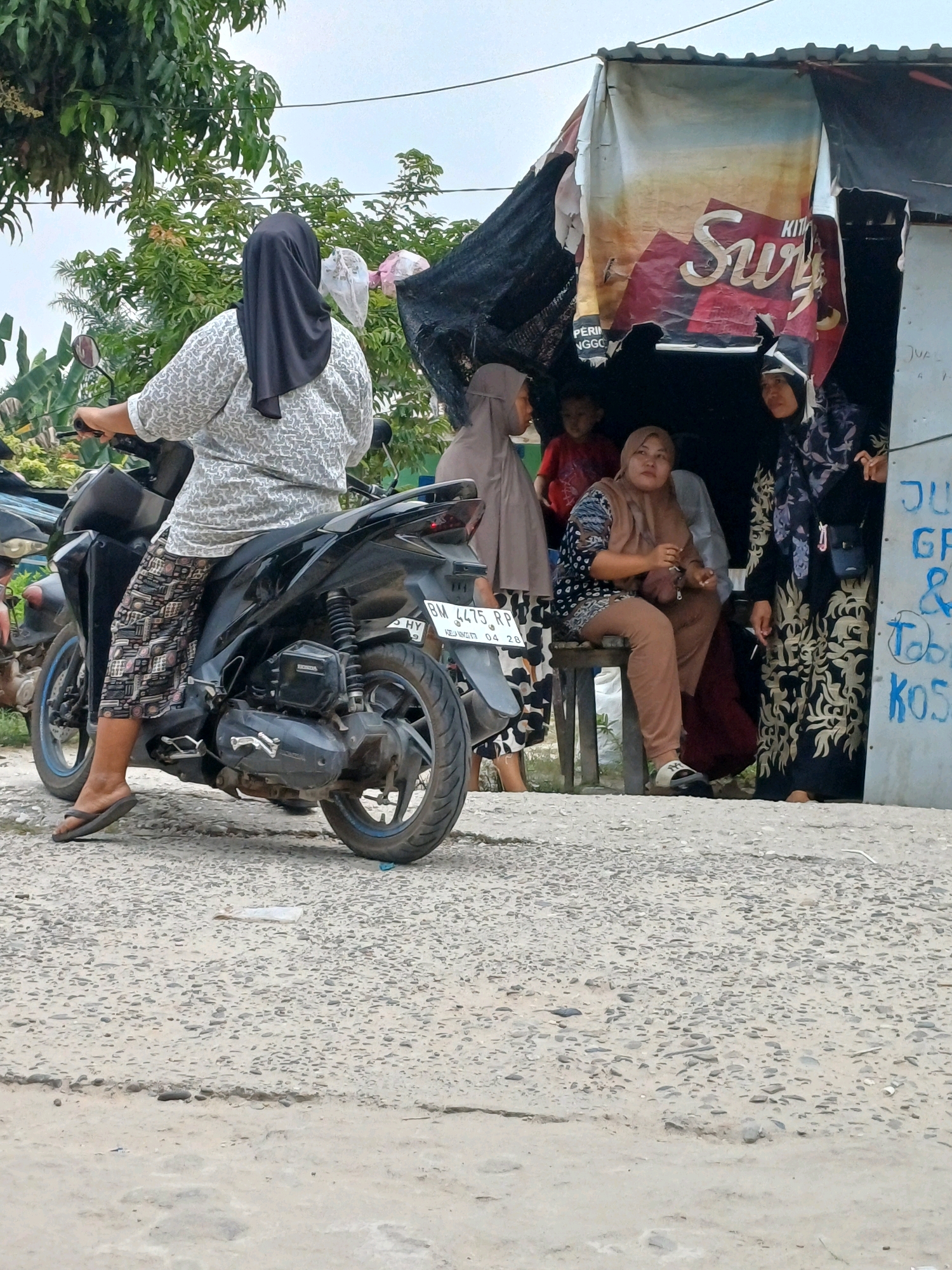 A group of people are gathered around a makeshift stall, possibly in a street market in Dumai City, Indonesia. In the foreground, a woman is seated on a black motorcycle, her back to the camera, wearing a patterned shirt and dark hijab. Her legs are crossed, and she wears flip-flops. The motorcycle's license plate reads "BM 4475 RP". Behind her, several other individuals are visible. One person, wearing a light brown hijab and pants, sits on a wooden stool. To their right, another person in a dark floral patterned hijab and dress stands. Further back, a child in a red shirt is partially visible. The stall itself is covered with a tattered tarp displaying an advertisement with the word "Sury" prominently featured. The ground is a rough, pebbled surface. The environment appears to be outdoors with some greenery and a cloudy sky. The stall has some writing on the side, including "JUAL" and what appears to be "KOST".
