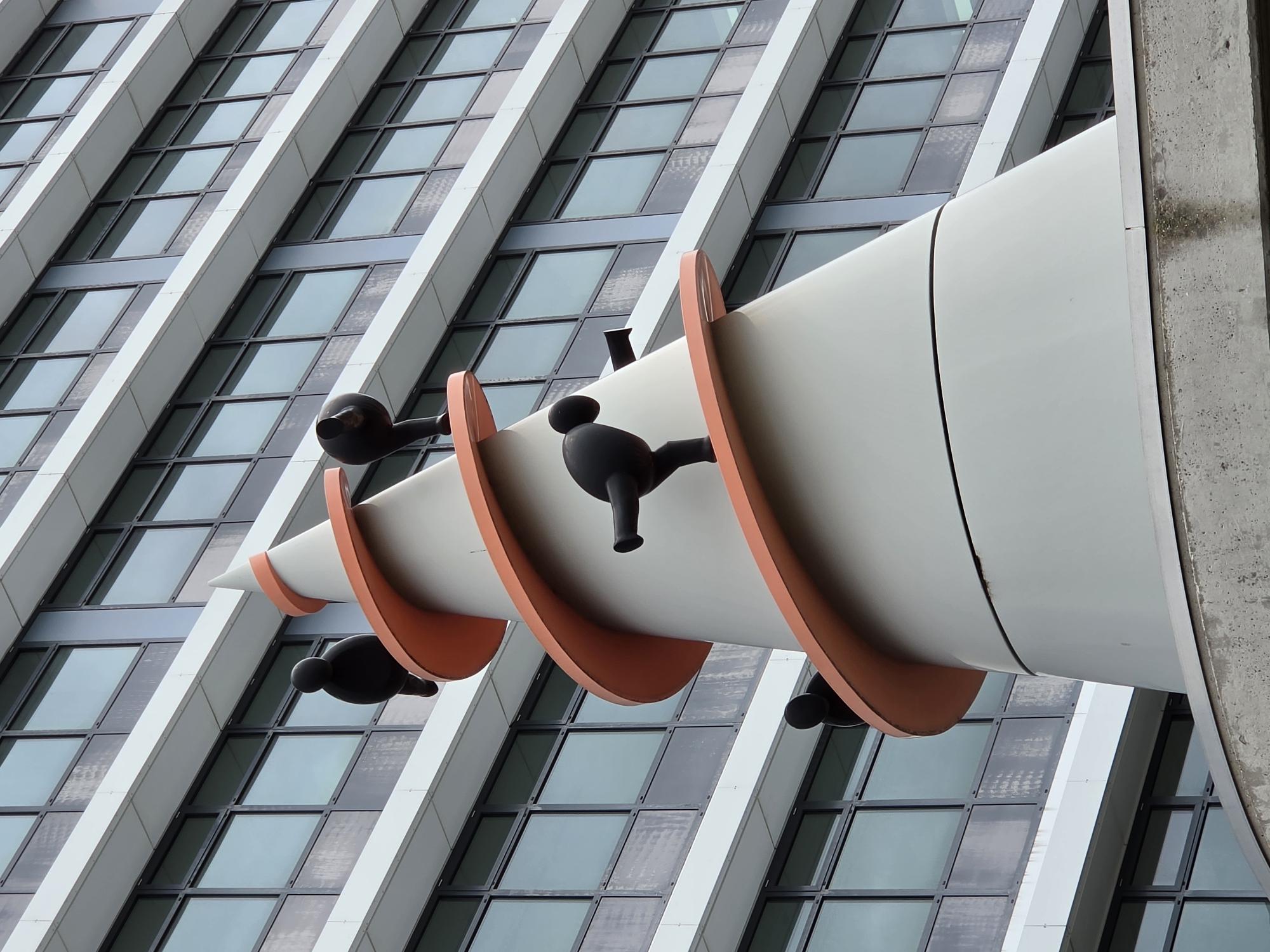 The image shows a close-up, low-angle shot of a conical sculpture with orange spiral accents, positioned in front of a modern building with numerous windows. Several black, abstract figures resembling stylized birds or humanoids are affixed to the sculpture at different levels. The sculpture appears to be made of a light gray material. The building behind it has a facade of white horizontal bands and rows of dark-framed windows. The scene is outdoors, with the sky reflected in the windows, suggesting a daylight setting. The sculpture is located at the museum Rijswijk, in Rijswijk, Netherlands.