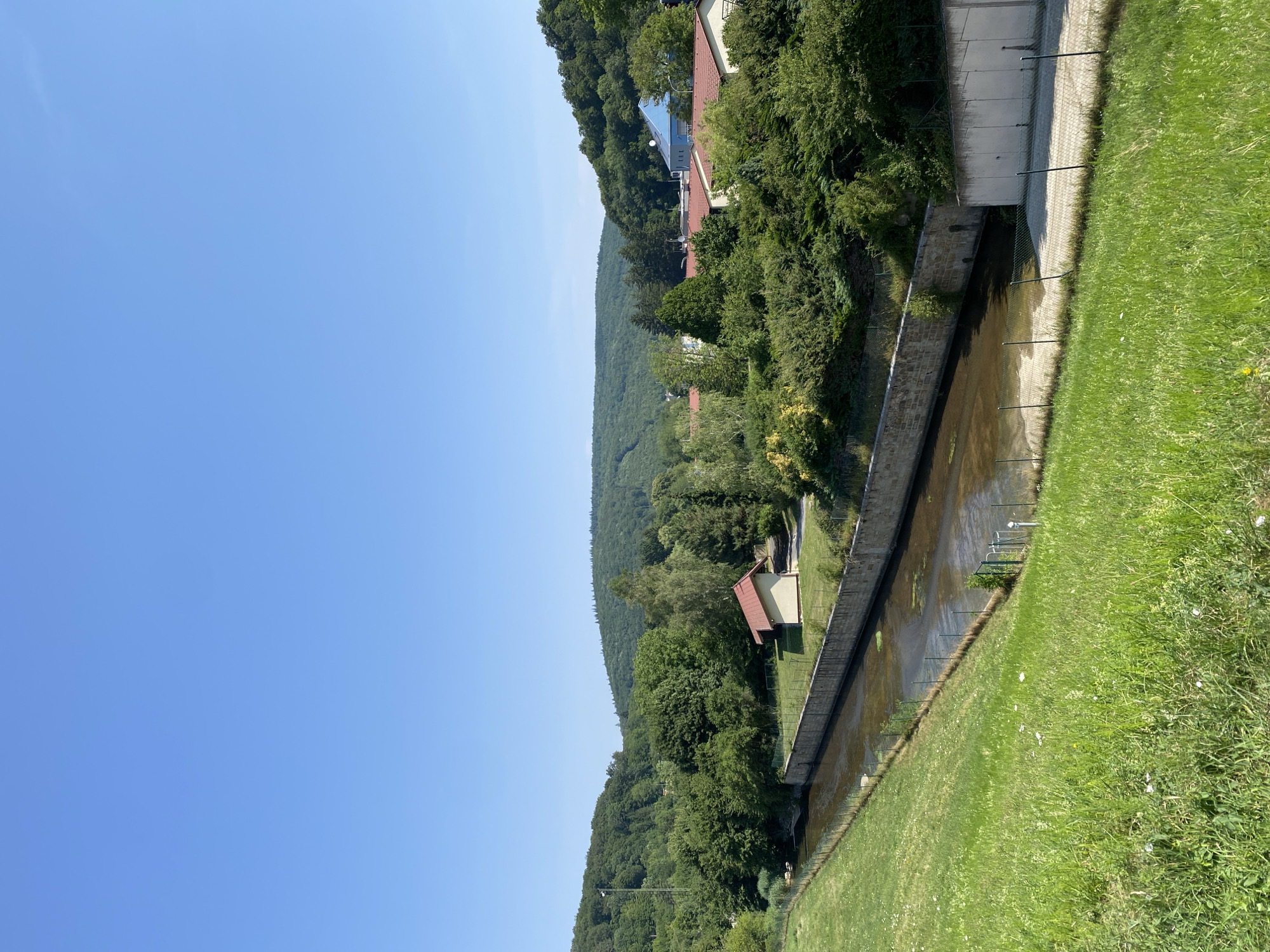 The image depicts a landscape scene in the city of Purkersdorf, Austria. In the foreground, a grassy slope descends towards a man-made channel with water flowing through it. The channel is bordered by a stone wall on one side and a fence. Beyond the channel, there is a small building with a red roof. Further in the background, lush green trees and hills dominate the scene, with several buildings with red tile roofs and one building with a blue roof visible amidst the foliage. The sky is clear and blue. The overall impression is one of a tranquil, rural environment on a sunny day.