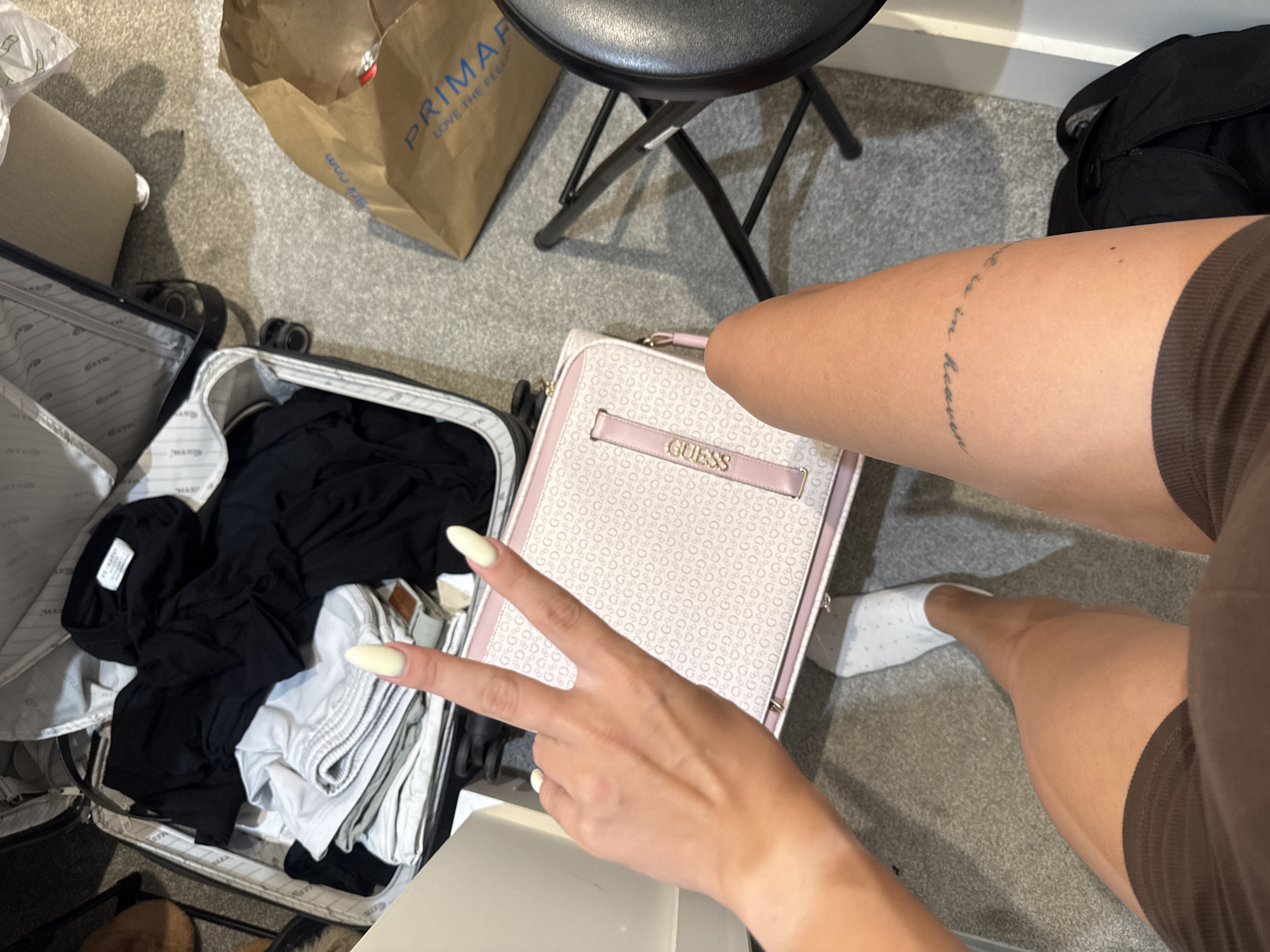 In this top-down perspective, a person's legs and hand are visible, indicating they are likely packing or unpacking a suitcase on a carpeted floor. The person is wearing brown shorts and white socks. Their left hand is raised, displaying a peace sign with painted nails. In front of them lies a light-colored bag with a repeating "G" pattern and a pink strap that reads "GUESS." To the left, an open suitcase is filled with black and white clothing. In the background, a brown paper shopping bag from Primark is on the floor, along with a black folding chair and part of a black backpack. The environment appears to be an indoor setting, possibly a bedroom or hotel room, in Throop, United Kingdom.