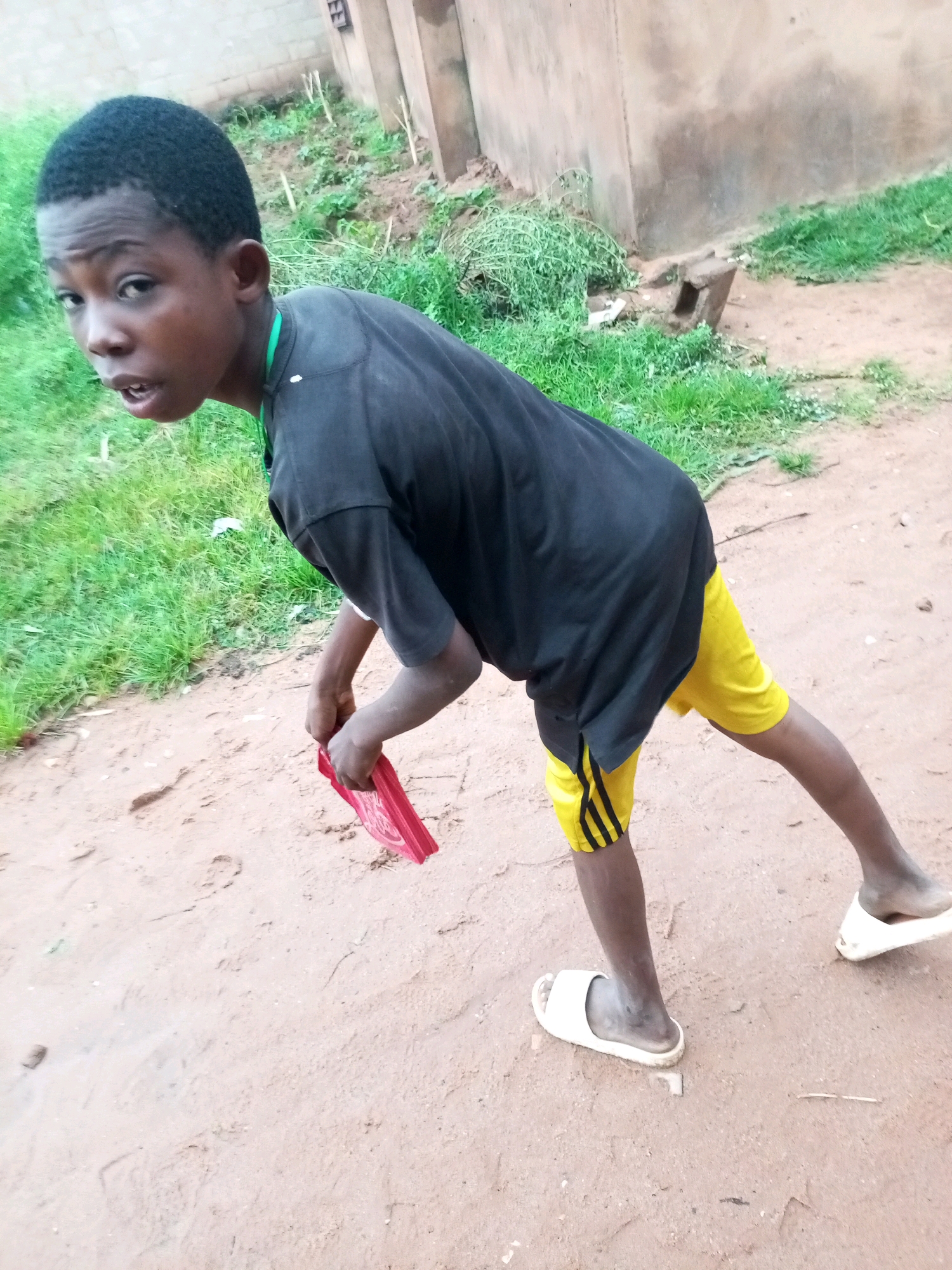 A young Black male, appearing to be in his early teens, stands on a dirt path outdoors. He is wearing a black t-shirt and yellow athletic shorts with black stripes. He is looking towards the camera with his mouth slightly open. His right hand is holding a folded red object. He is wearing white slide sandals. The background shows a light-colored building with some greenery and dirt. The scene appears to be in a residential area in Gombe, Nigeria.