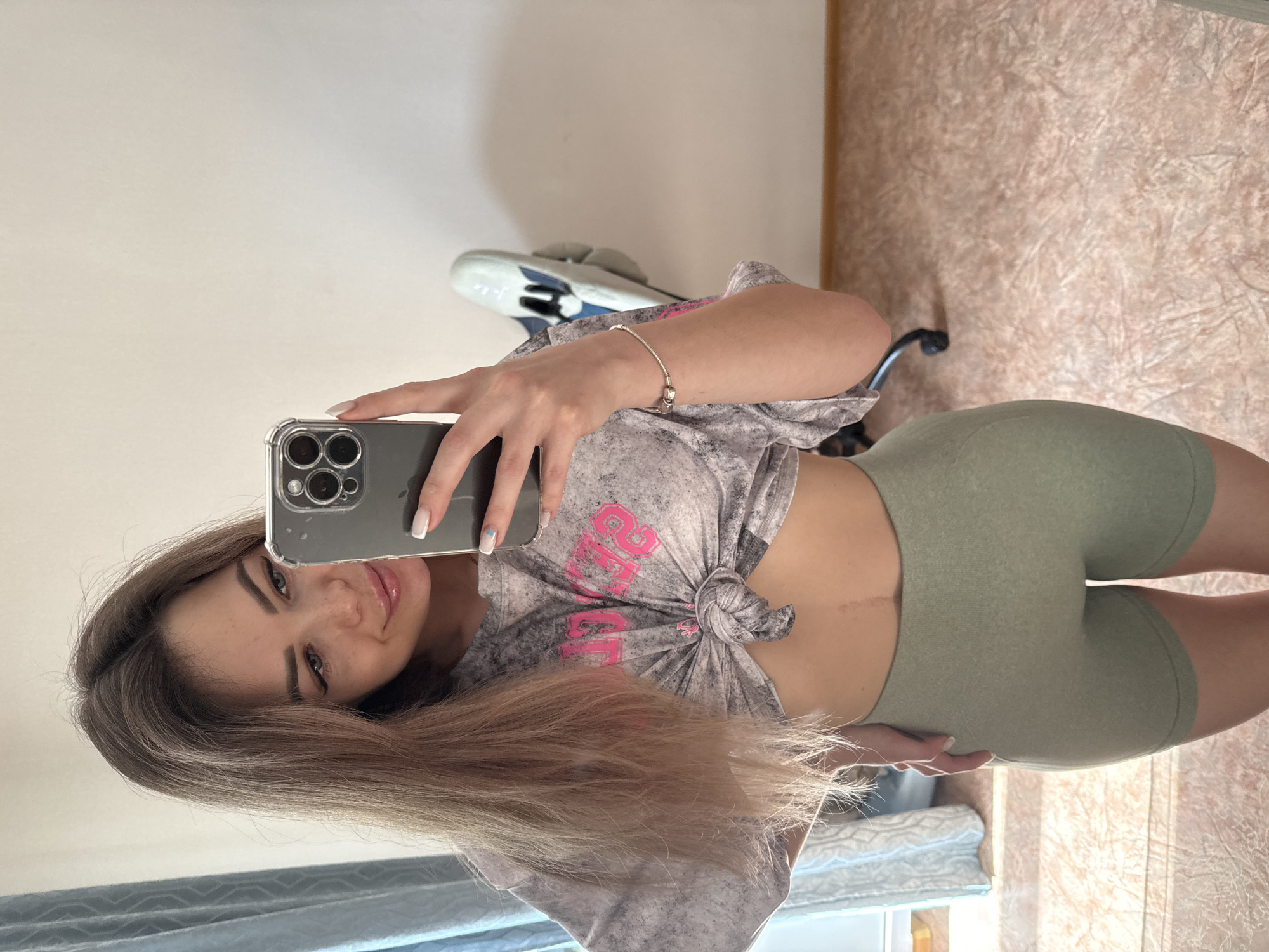 A female with long light brown hair is holding a phone to take a selfie. She's wearing a gray and pink t-shirt tied in a knot, and light green biker shorts. The background includes a wall, a floor, and a curtain. There is also a pair of sandals and a chair visible in the background. Location: Unknown, Unknown.