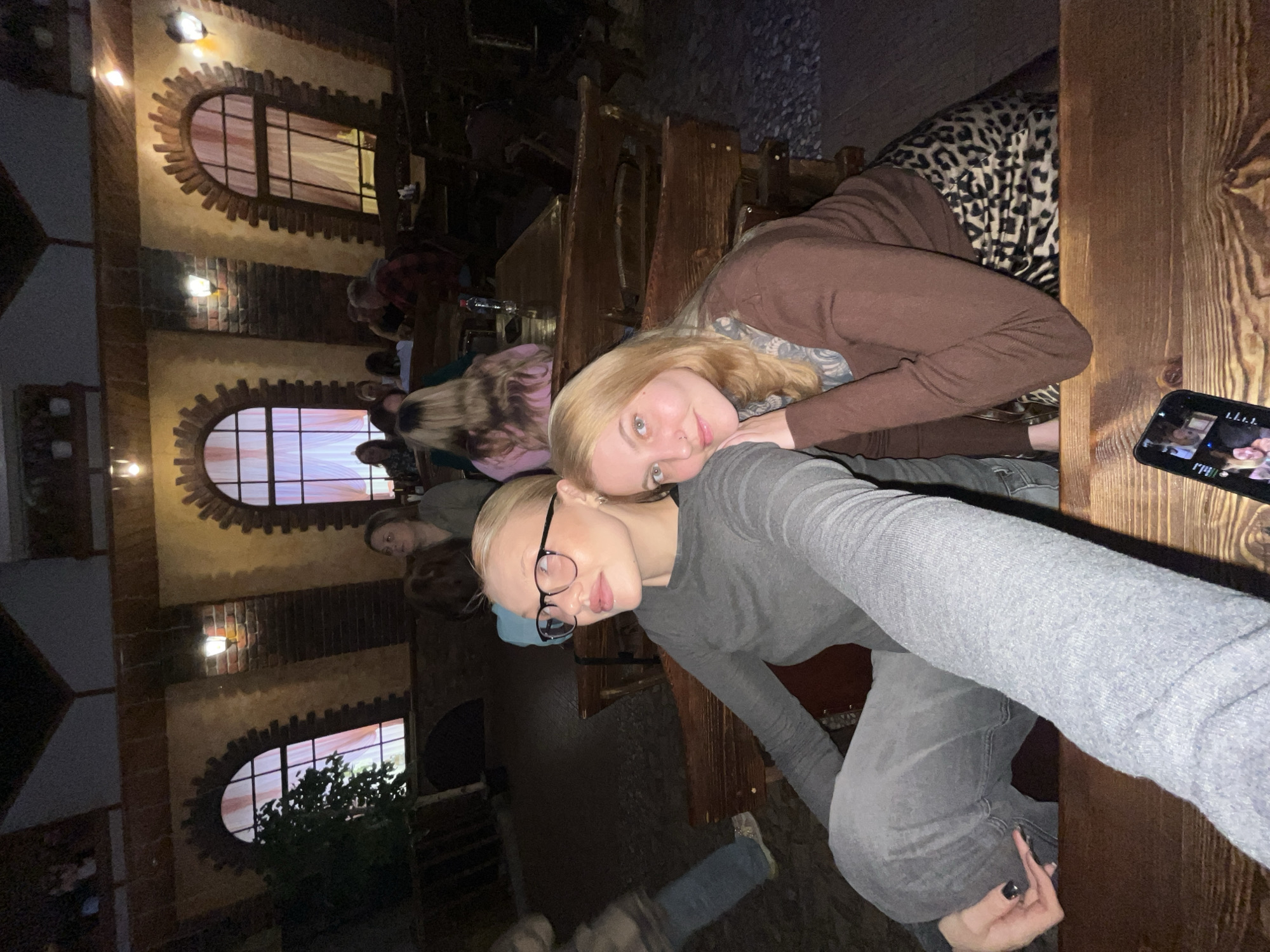 Two young women are posing for a selfie indoors. One woman is wearing a gray long-sleeved shirt and glasses, and the other is wearing a brown outfit and a patterned scarf. They appear to be sitting at a wooden table. A cell phone is on the table, displaying a video. Other people and tables are visible in the background, along with arched windows. The location appears to be a restaurant or similar establishment in Chaikovsky, Russia, as indicated by the architectural style and interior design.