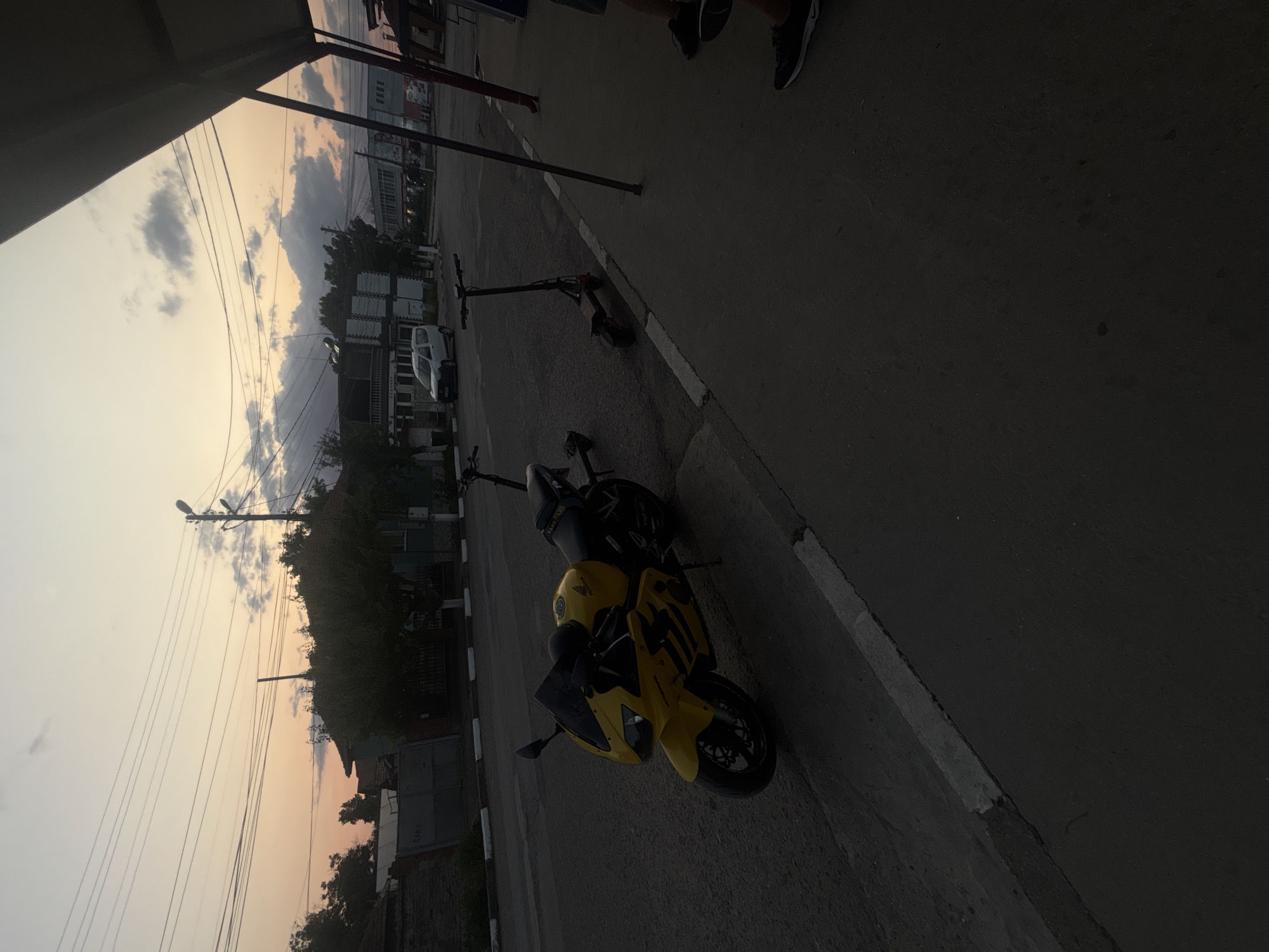 The image captures a street scene in Marten, Bulgaria, during what appears to be dusk, evidenced by the soft, warm hues in the sky and the dim lighting. In the foreground, a bright yellow sport motorcycle is parked on the asphalt next to a sidewalk. Beside the motorcycle, two electric scooters are also visible. The street is lined with trees and various buildings, including what looks like a white van parked on the opposite side of the road. Power lines crisscross the sky, and a street lamp is present. The overall atmosphere suggests a quiet, late afternoon or early evening setting in a residential or commercial area of the town.