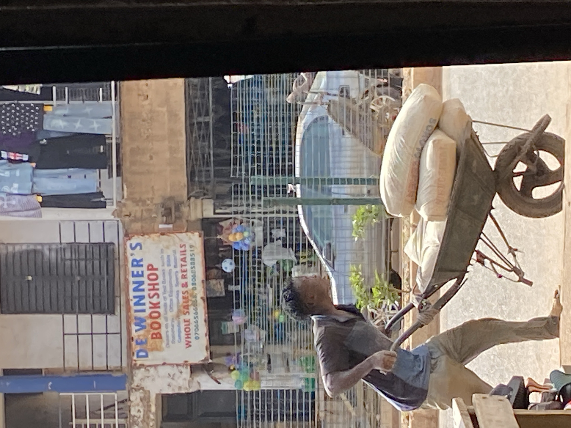 In Bukuru, Nigeria, a man is pushing a wheelbarrow loaded with sacks of cement. He is wearing a dark t-shirt with a blue panel on the front and light brown trousers. His left foot is bare. The wheelbarrow has a single wheel and is made of dark metal. Behind the man, a silver car is parked. A shop sign for "DE WINNER'S BOOKSHOP" is visible, displaying text about "WHOLE SALES & RETAILS" and phone numbers. The shop appears to sell books, stationery, and possibly other items. In the background, there are multiple shops with clothing displayed on a balcony. The scene is outdoors, with sunlight illuminating the area.