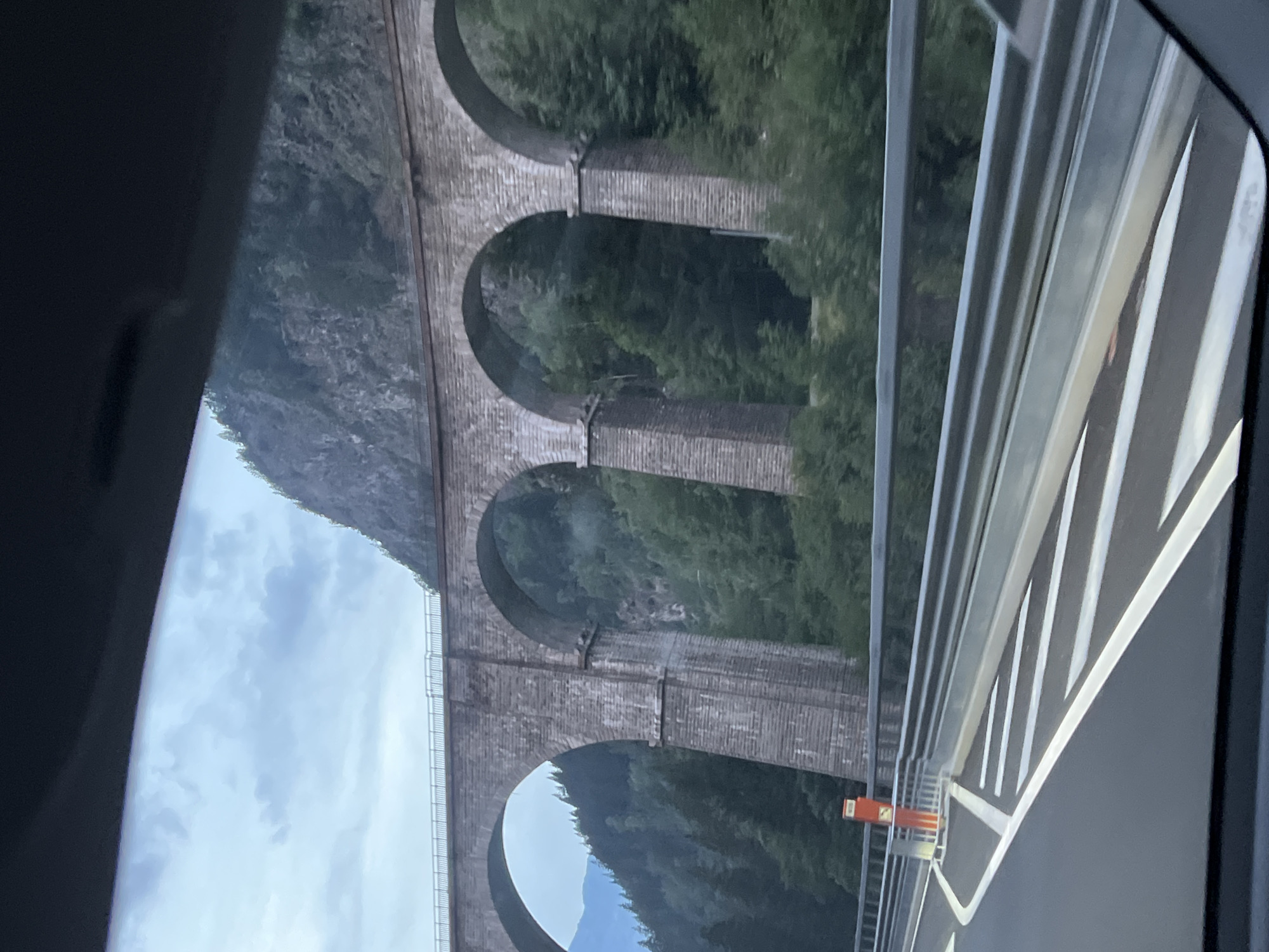The image captures a stone viaduct with multiple arches, likely a railway bridge, spanning across a lush, green valley. The viaduct is constructed from large, rough-hewn stone blocks and features robust, tapering pillars supporting the arches. The top of the viaduct appears to have a railing, and what looks like railway tracks can be seen. The surrounding environment is densely forested with tall, dark green trees. A steep, rocky mountain slope rises behind the viaduct, with a cloudy sky overhead. In the foreground, a modern road with white lane markings and a metal guardrail is visible, suggesting the photograph was taken from a vehicle on a road below or adjacent to the viaduct. An orange traffic marker is present on the side of the road. The location is identified as a viaduct in Les Houches, France.