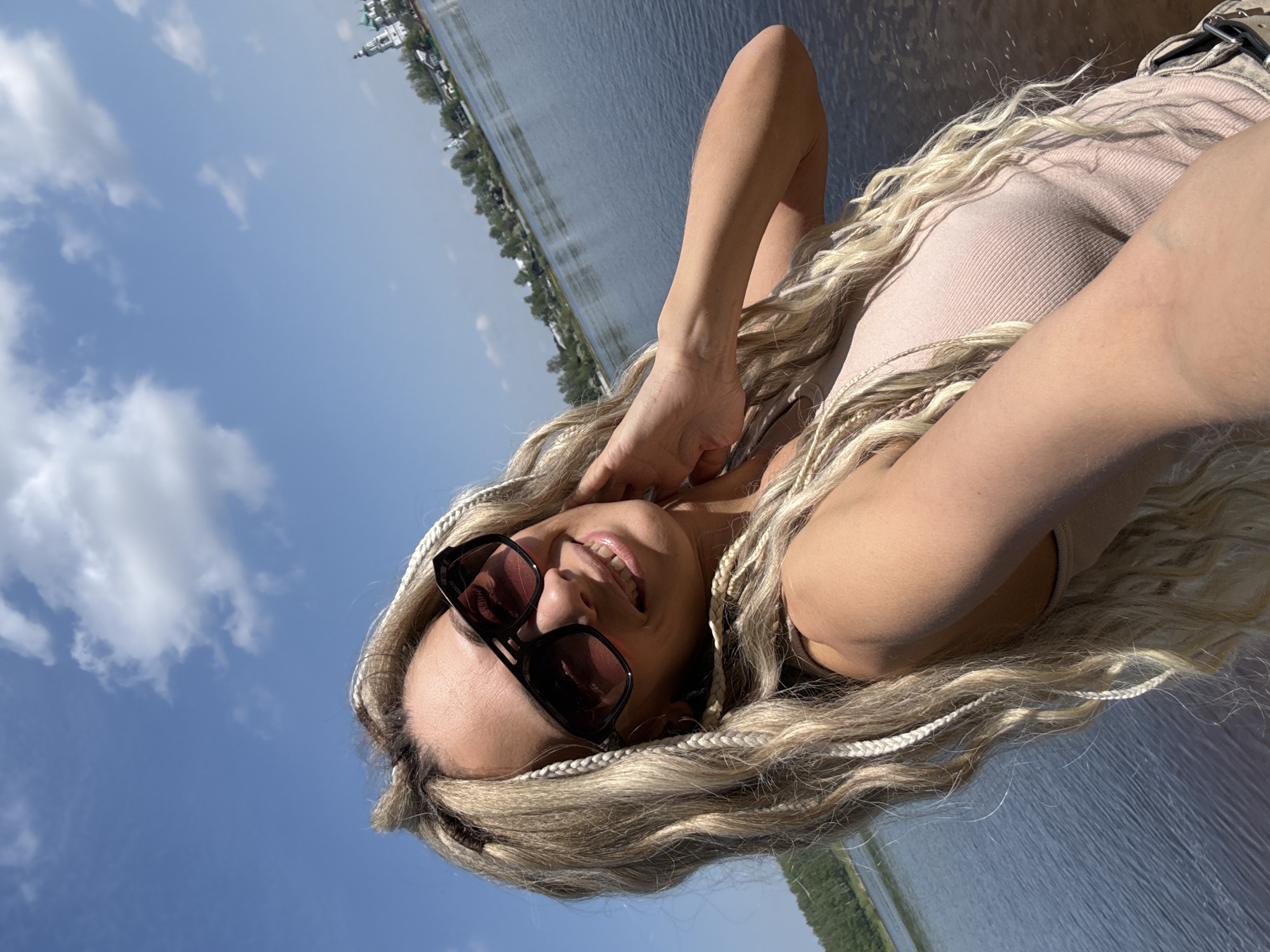 A selfie shot shows a woman with blond hair and white braids lying down on what appears to be a wooden surface next to a body of water. She is wearing sunglasses and a light-colored tank top. The background features the body of water with trees lining the opposite bank, and a cloudy blue sky above. The city is not discernible.