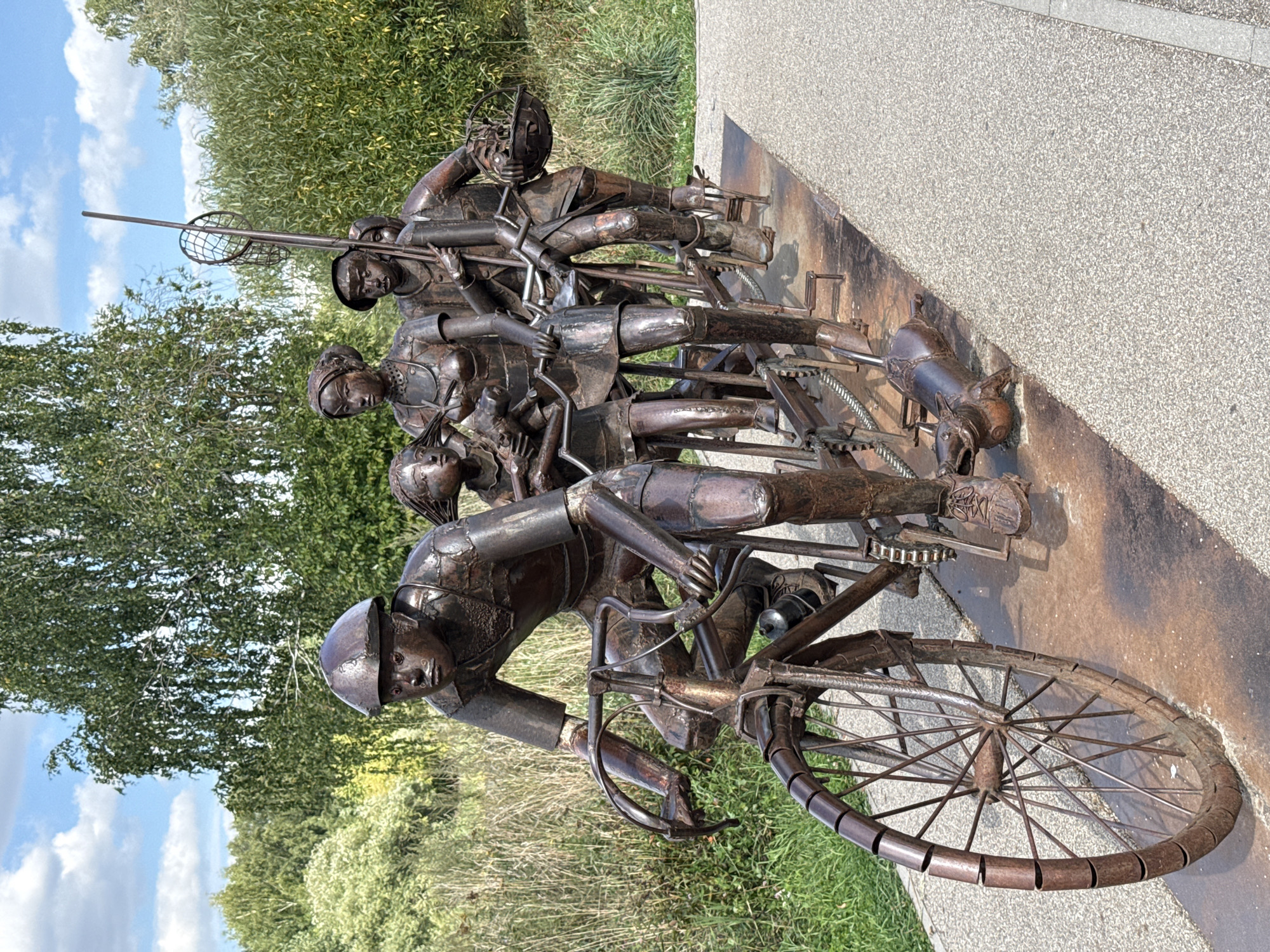 A full shot of a bronze sculpture depicts a family of five riding a tandem bicycle, with a pig trotting alongside. The sculpture is in a park-like setting with trees and grass in the background. The sculpture is made of weathered bronze, with visible rivets and seams. The family is dressed in early 20th-century attire. The father, wearing a helmet, is pedaling the front of the bicycle. His wife sits behind him, holding a baby. A young boy sits behind his mother, holding a fishing net on a pole. Another child sits behind the boy, holding a basket. The pig is depicted with its mouth open, as if barking. The sculpture is placed on a concrete path, which is adjacent to a grassy area. The sky is blue with scattered clouds.