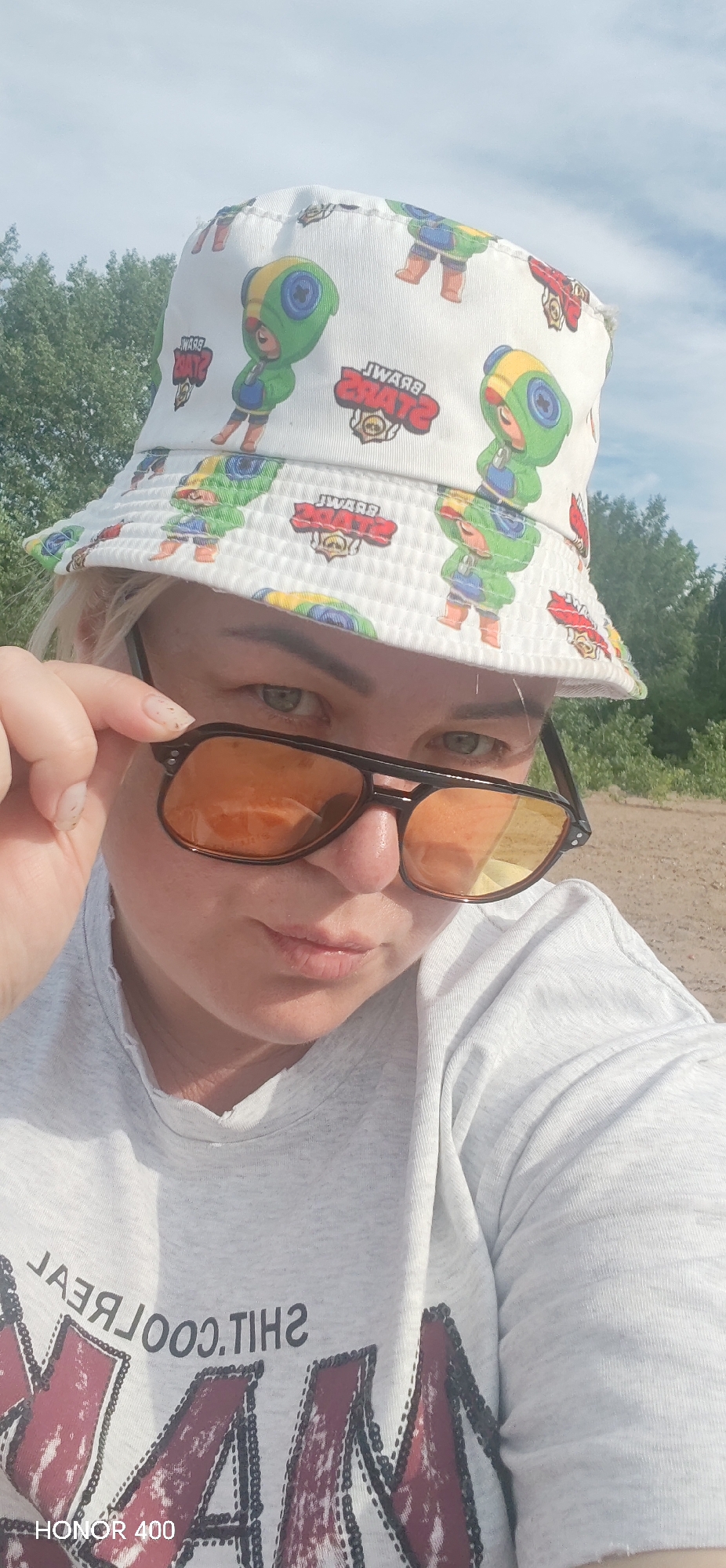 A woman is featured in a close-up selfie taken outdoors in Noviy, Russia, on a bright, sunny day. She has blonde hair and is wearing a white bucket hat with colorful cartoon characters and text, along with a light grey t-shirt. The characters on the hat are identifiable as Leon from the mobile game "Brawl Stars," and the mirrored text "BRAWL STARS" is also visible. She is seen adjusting dark-framed, orange-tinted sunglasses, pulling them down slightly to reveal her light-colored eyes, which are looking directly at the camera with a neutral expression.
The background shows green trees and a clear blue sky, indicating an outdoor, natural setting. The ground appears light and somewhat sandy. On her t-shirt, mirrored text is visible, reading "THIS REAL" as part of a larger, partially obscured design. A "HONOR 400" watermark is present in the bottom left corner of the image.