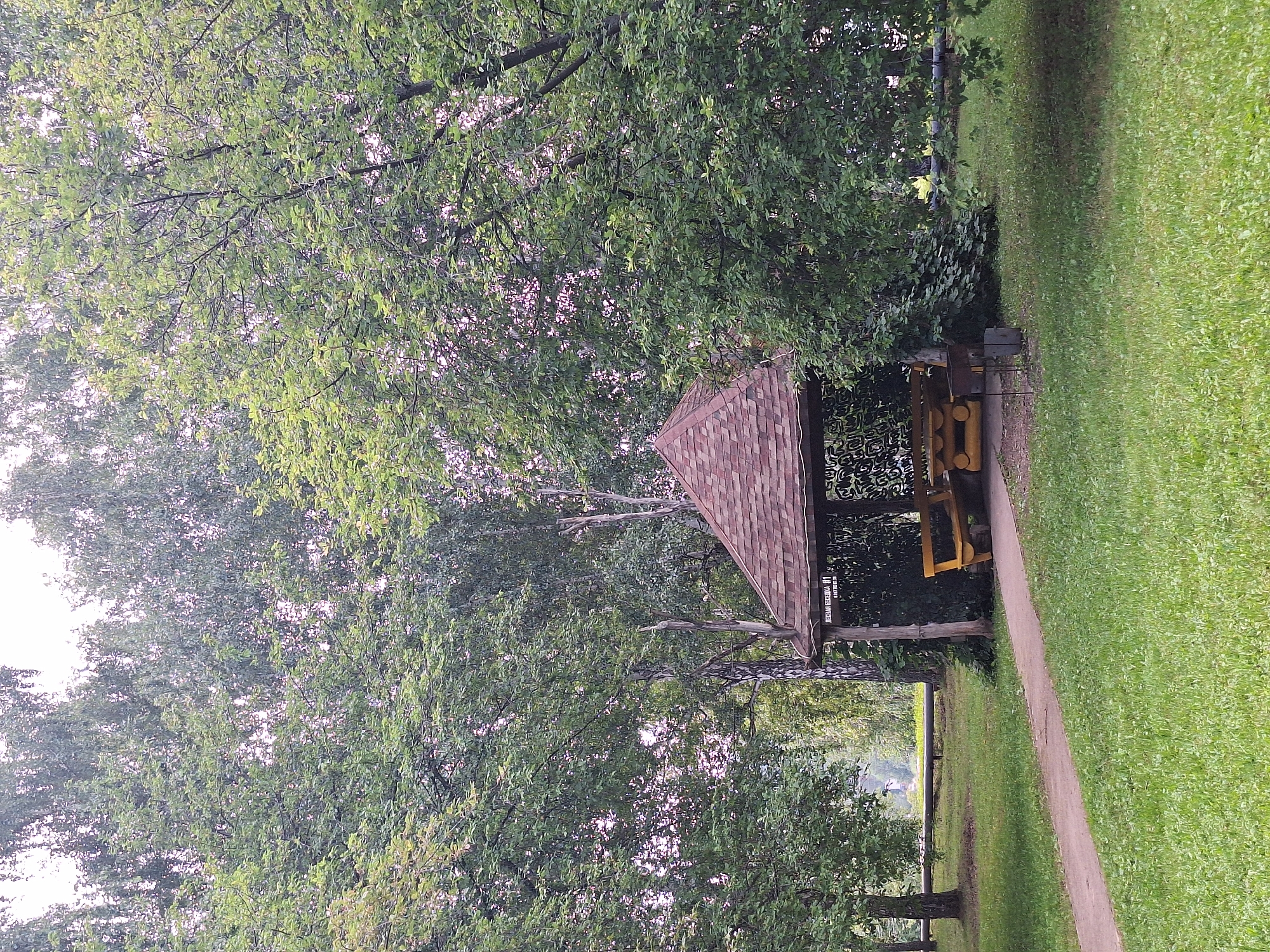 A wooden gazebo with a brown, shingled roof is situated in a grassy park, surrounded by lush green trees. Inside the gazebo, there is a wooden picnic table and benches. A paved walkway leads up to the gazebo. In the background, a metal pipe runs horizontally across the scene, and a distant building can be seen. The sky is overcast. The sign on the gazebo reads "ЛЕСНАЯ БЕСЕДКА №1".