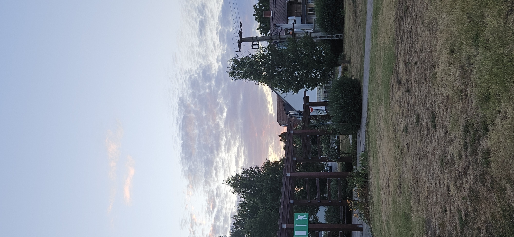The image depicts an outdoor scene at dusk in the city of Hévíz, Hungary. In the foreground, a patch of dry, brown grass is visible, with a paved walkway curving through it. Behind the grass, a wooden pergola structure stands, with a green sign featuring an exit symbol and a running figure attached to its left side. The pergola casts shadows on the ground. Beyond the pergola, several trees with dark green foliage are present, some taller than others. To the right, a telephone pole with wires and an electrical transformer is situated. In the background, residential buildings with tiled roofs are visible, one with white siding and a reddish-brown roof, and another with a terracotta-colored roof. The sky above is a pale blue, with scattered clouds tinged with pink and purple from the setting sun. The overall impression is a calm, suburban environment during twilight.