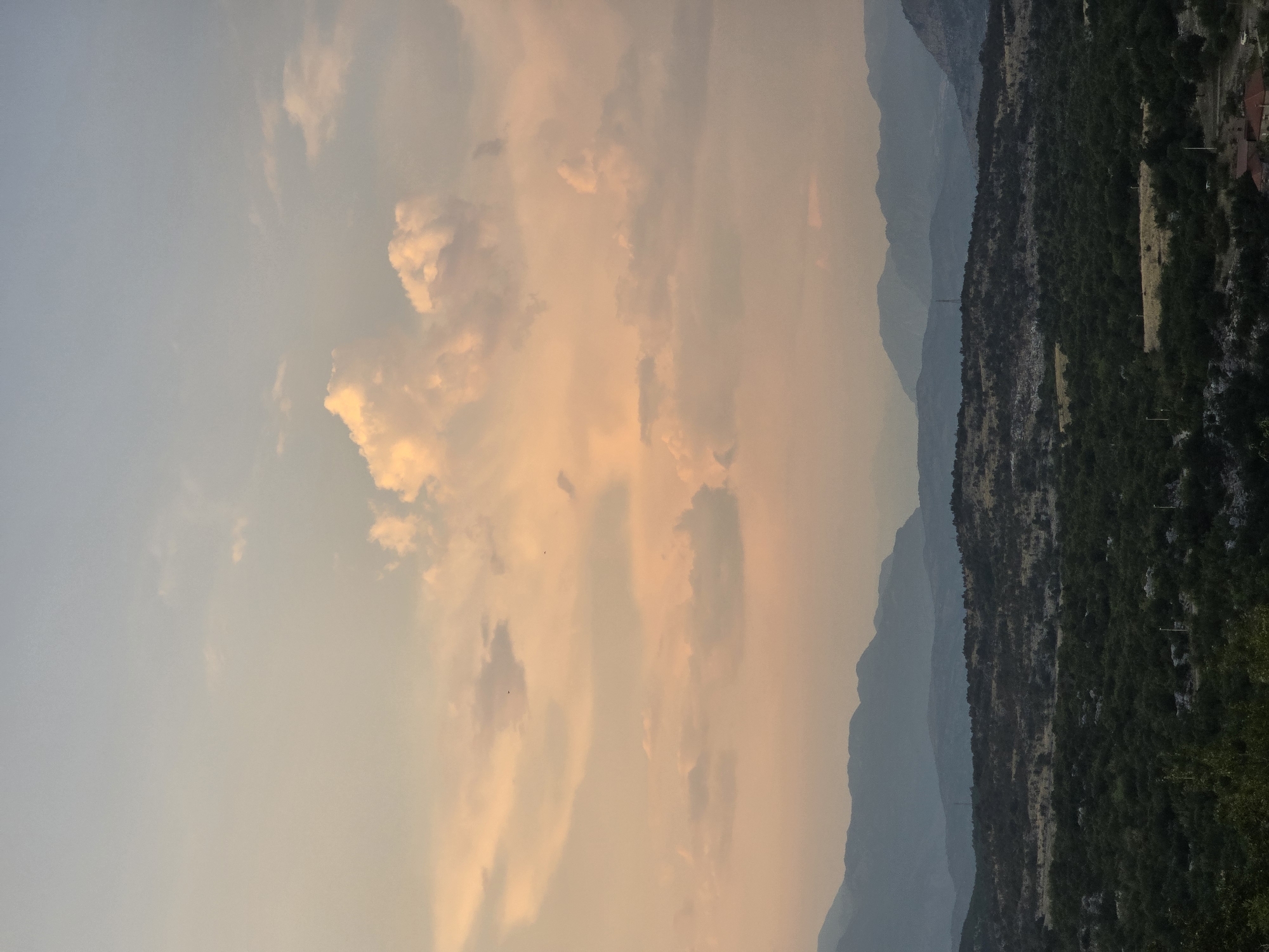 The image depicts a landscape view from Medun, Montenegro, during what appears to be sunset or early evening. The sky is filled with soft, warm-toned clouds in shades of pink, orange, and light blue, suggesting the golden hour. Below the sky, a series of layered mountains recedes into the distance, rendered in muted blues and grays due to atmospheric perspective. The foreground features a dense, dark green forest or scrubland covering undulating hills. Scattered throughout this lower area are patches of lighter vegetation and some indistinct structures or poles. The overall atmosphere is tranquil and natural, with no visible people or animals. The scene captures a serene natural vista with distinct mountain ranges and lush vegetation.