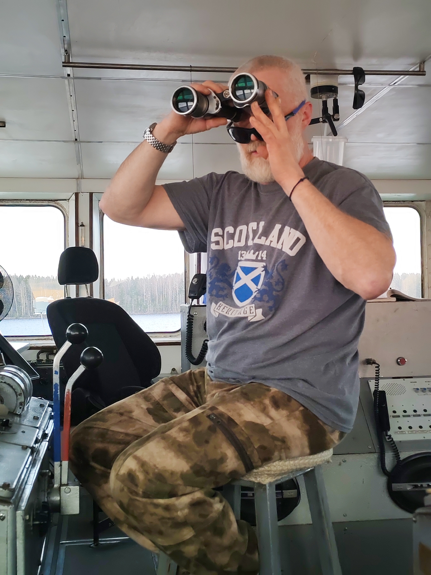 A man with a white beard and gray hair is sitting on a stool inside a boat or ship, looking through binoculars. He is wearing a gray t-shirt with "Scotland" printed on it and camouflage pants. A watch is visible on his left wrist, and sunglasses are perched on his head. He has a bracelet on his right wrist. The setting appears to be the interior of a ship's bridge or control room, with windows revealing a body of water and trees in the background. There is navigational equipment and controls visible, including levers and a telephone-like device. The exact location in Anisimovo, Russia, cannot be determined from the image.