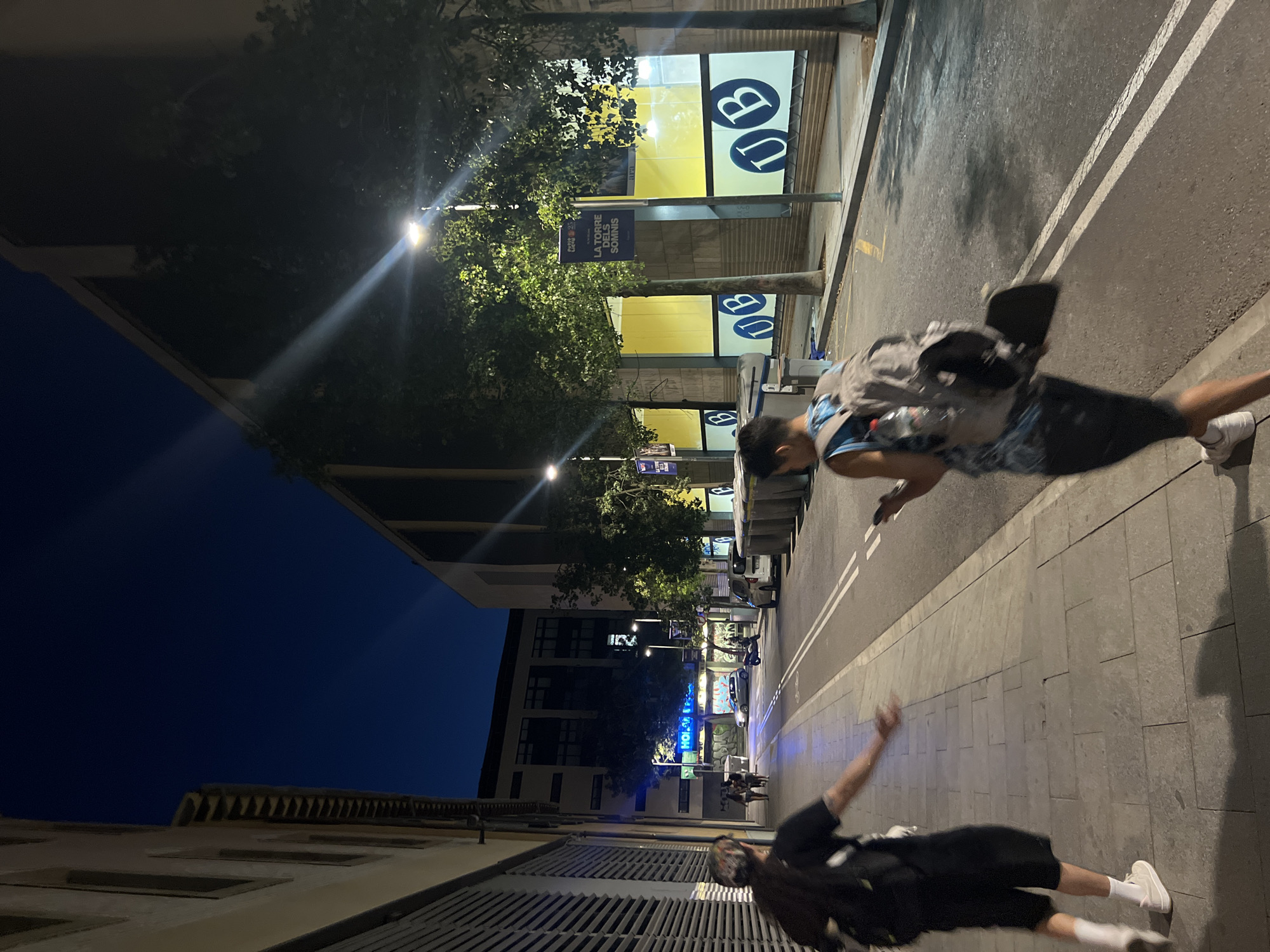 The image depicts a night scene on a city street in Barcelona, Spain. In the foreground, two individuals are walking away from the camera. The person on the left is wearing a black t-shirt, black shorts, white socks, and white sneakers, carrying a black backpack. The person on the right is wearing a blue and white patterned tank top, dark shorts, and white sneakers, also carrying a backpack with a skateboard attached. The street is paved with grey tiles on the sidewalk and asphalt on the road, marked with double white lines.
The environment includes a building on the left with a ribbed facade. On the right, there is a building with large windows covered in yellow material, featuring the blue "UB" logo repeated multiple times. A street sign that reads "LA TORRE DELS SOMNIS" is visible on a pole in front of the building. Trees line the sidewalk, illuminated by streetlights. Several large, grey trash bins are placed along the sidewalk. In the background, other buildings and streetlights are visible, and a silver car and a scooter are parked on the street. The sky is dark blue, indicating nighttime.